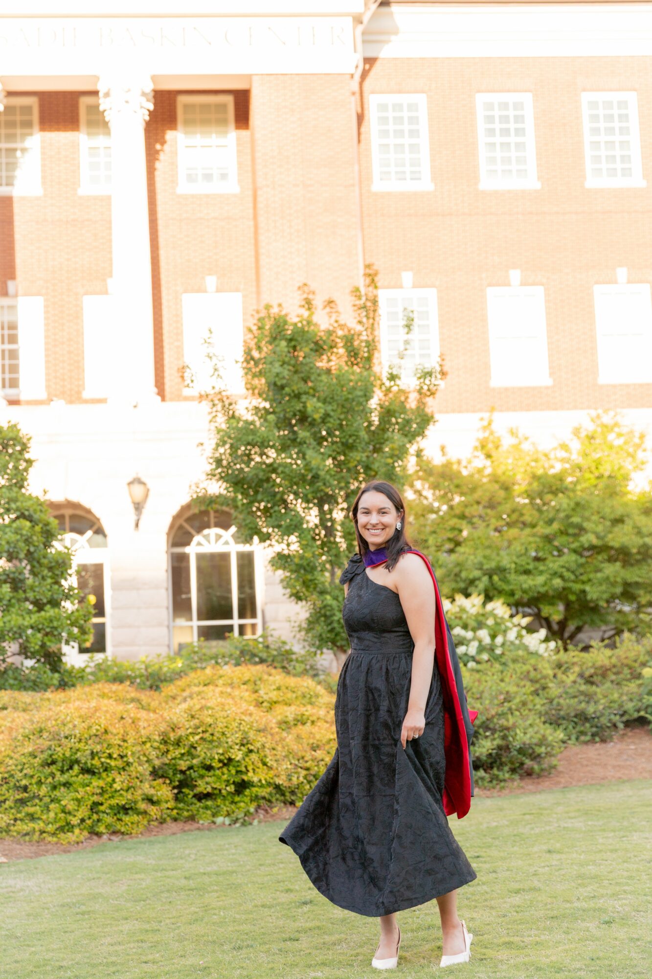 Woman in a black dress and red cape standing on grass with building and trees in background.