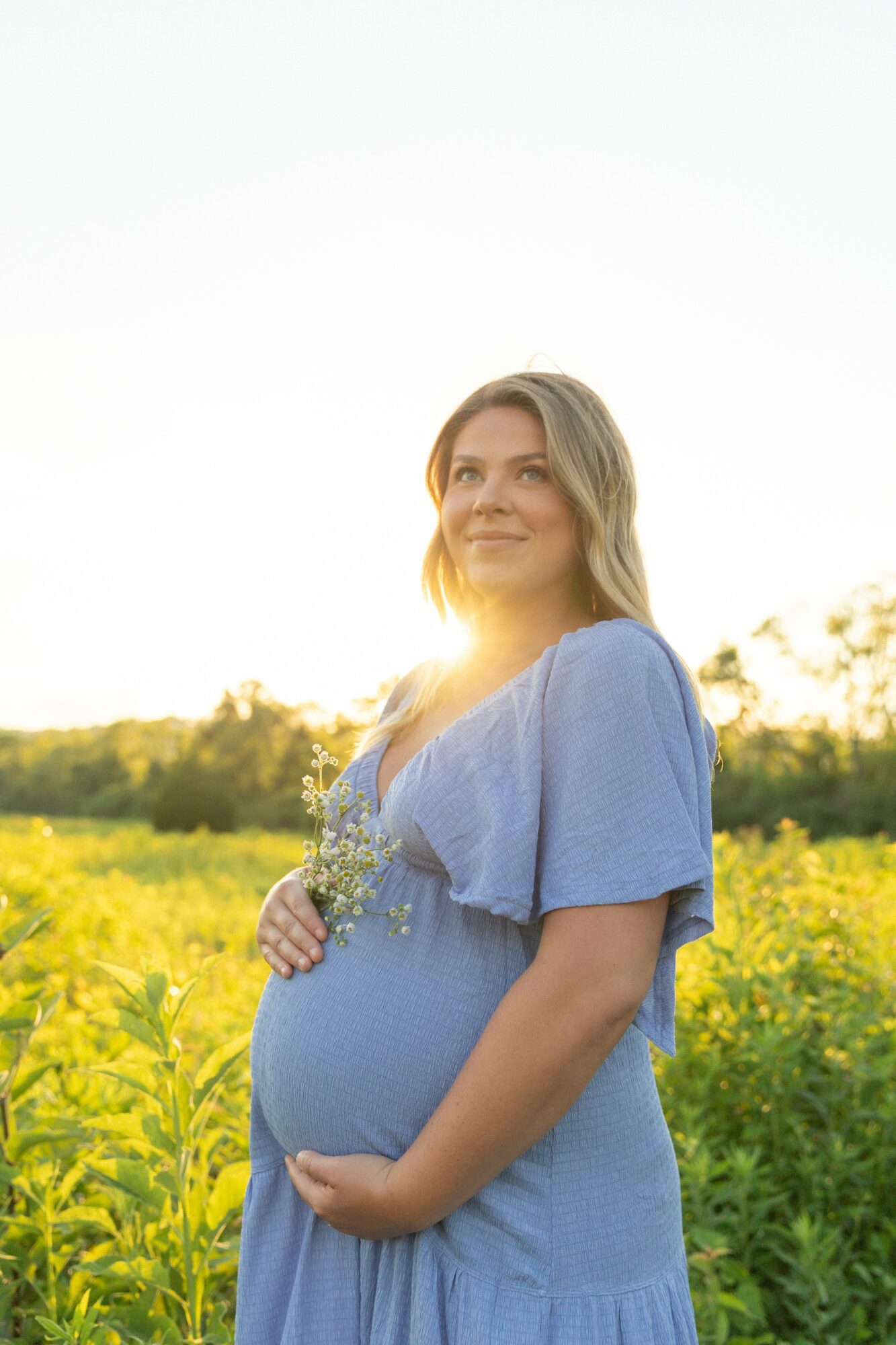 Pregnant woman in a blue dress standing in a field of yellow flowers during sunset, smiling gently, hands on her belly.