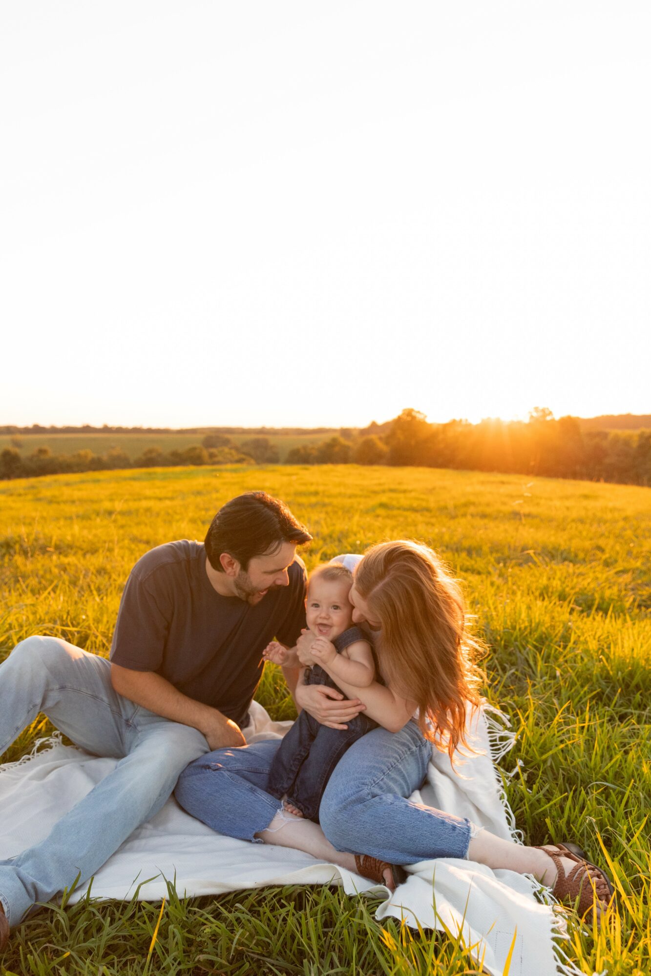Family sitting on a blanket in a field during sunset, with a man, woman, and child interacting happily.