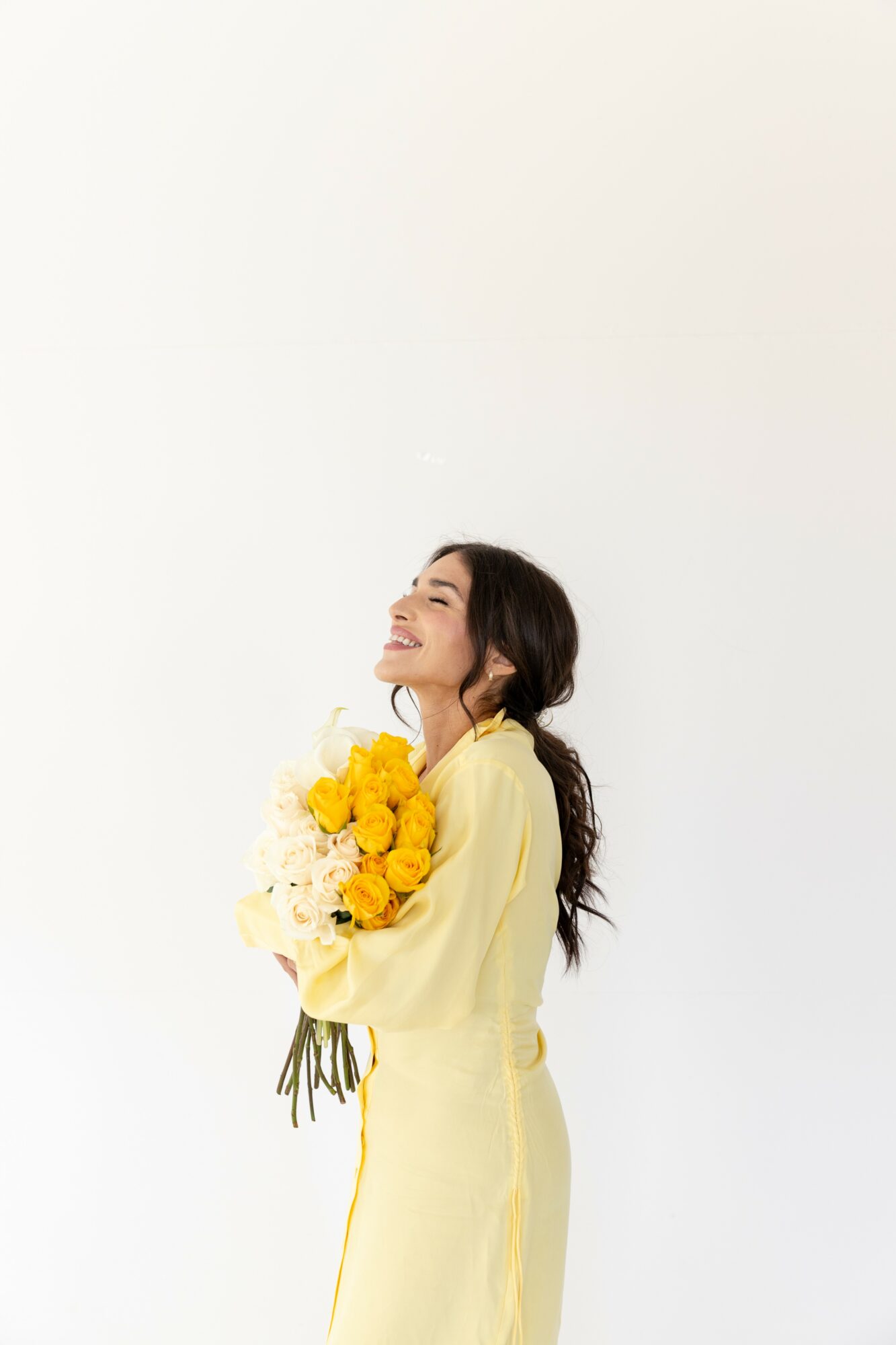 Woman in yellow dress holding a bouquet of yellow flowers, smiling with eyes closed, against a plain white background.