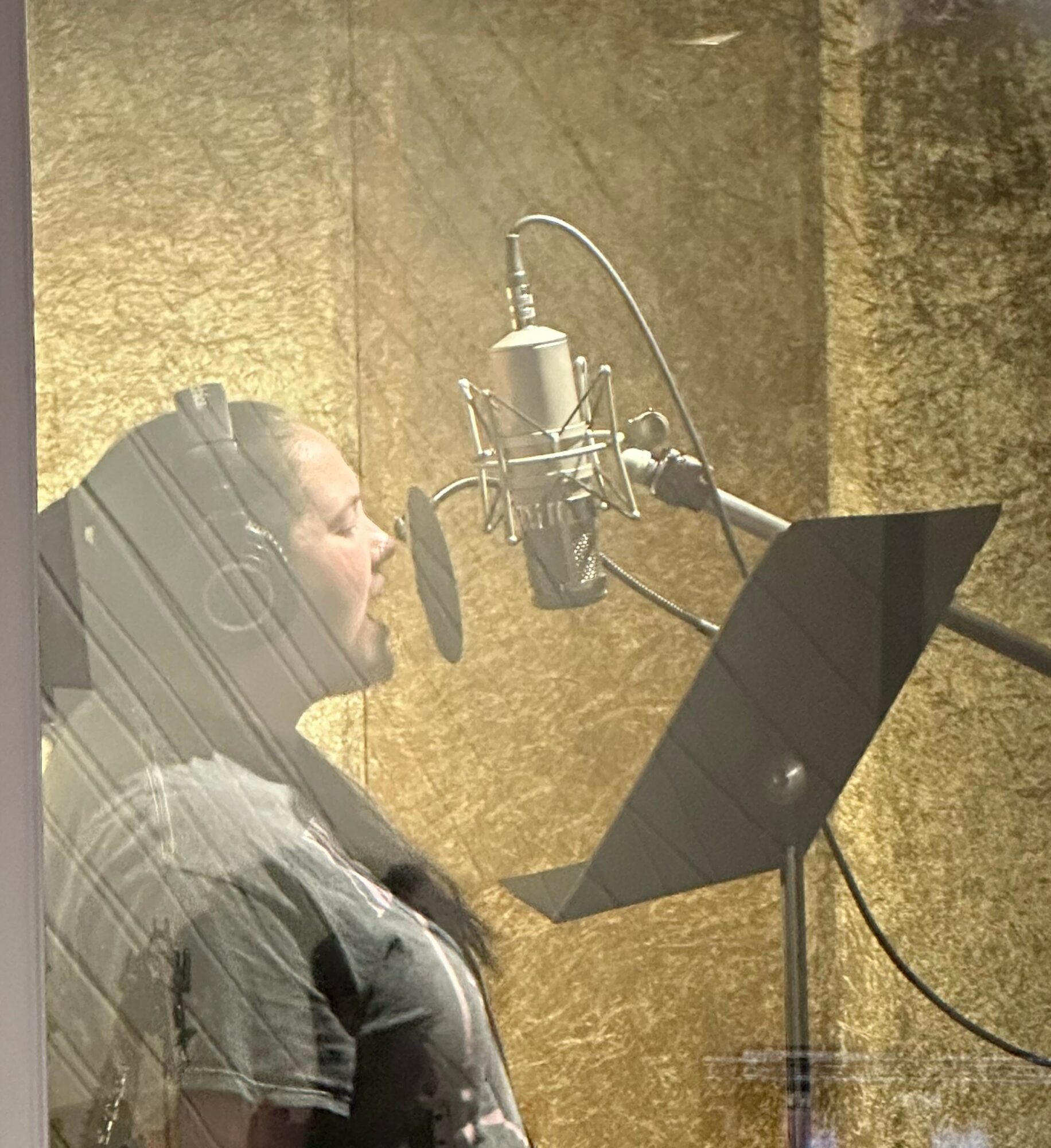 Woman singing into a microphone in a recording studio with a music stand and gold-colored wall background.