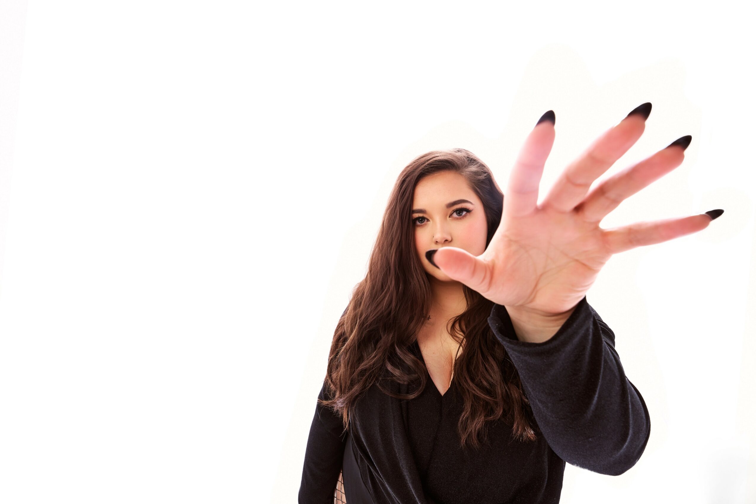 Young woman with long dark hair extending her hand towards the camera, blocking her face, wearing a black top.