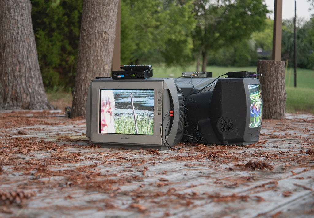 A video camera on the ground outdoors captures trees and grass, with a screen showing a view of the scene.