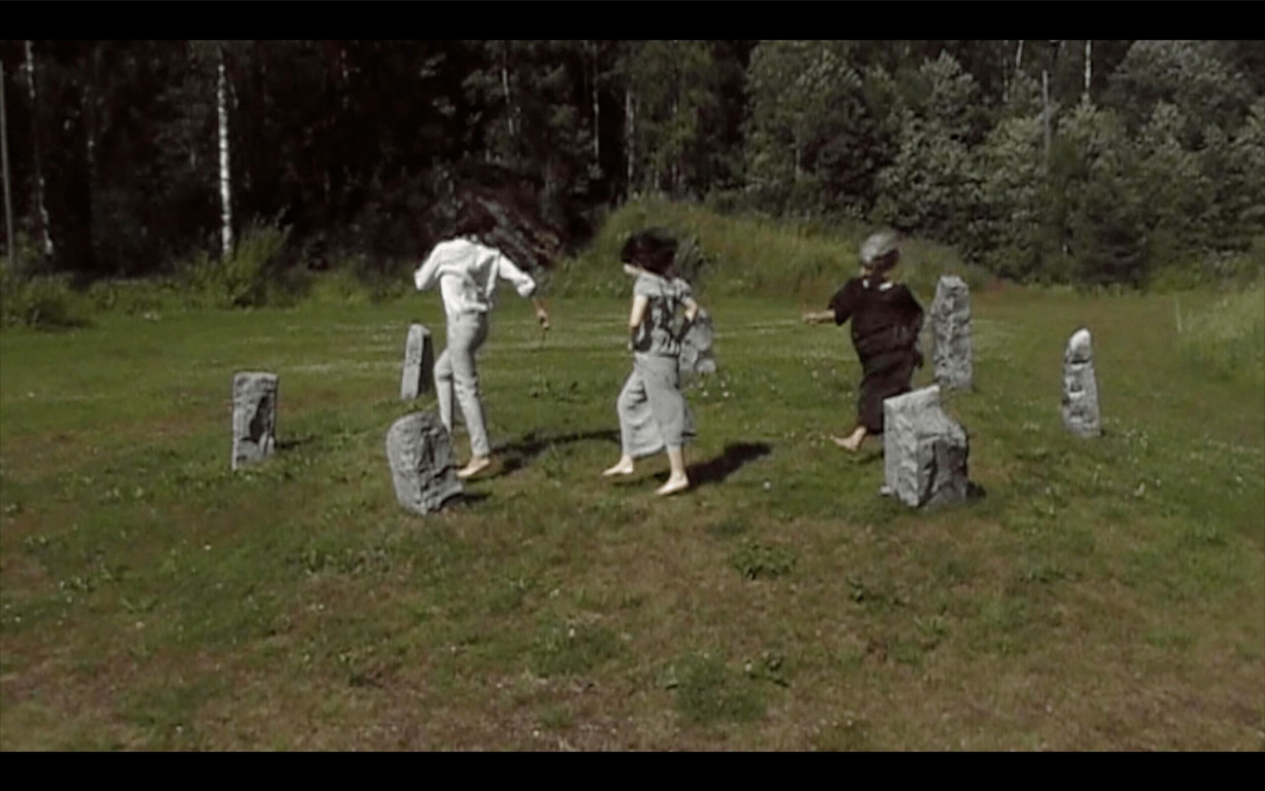 Three people are walking among small stone markers in a grassy area with trees in the background.