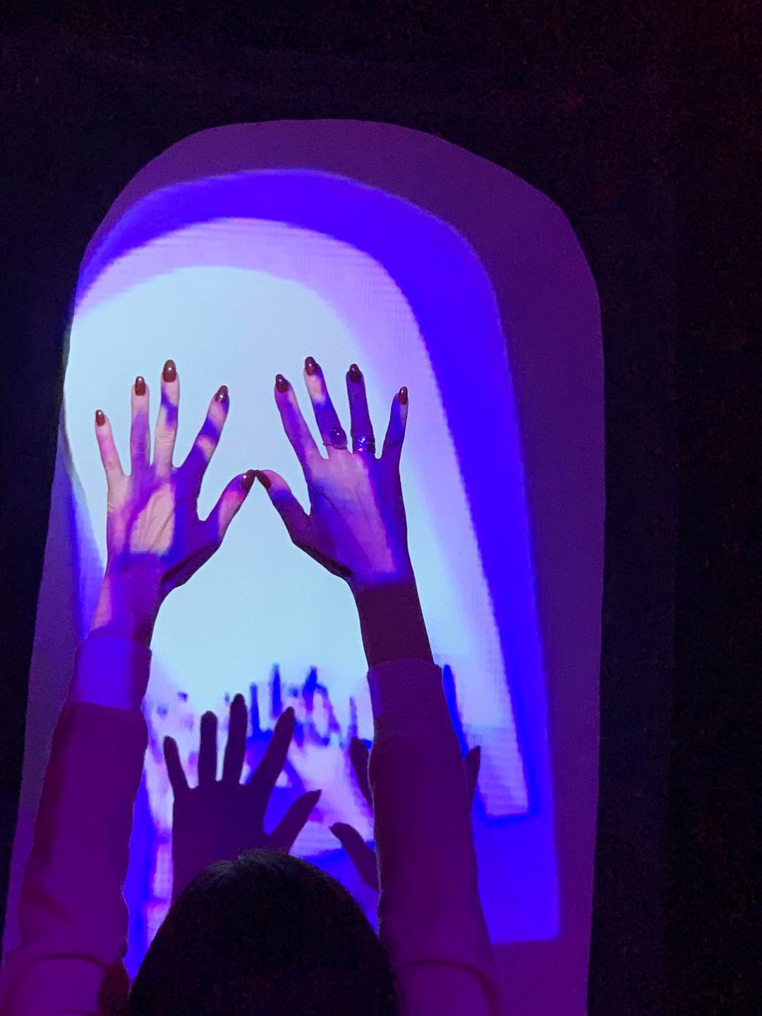 Two hands reaching towards a colorful, glowing rainbow projection on a dark background.