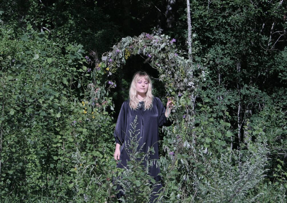 Young woman standing in front of a floral arch outdoors surrounded by greenery.