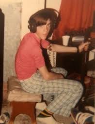 Person with shoulder-length hair wearing a pink shirt and plaid pants, sitting on a bench, reaching for a remote control.