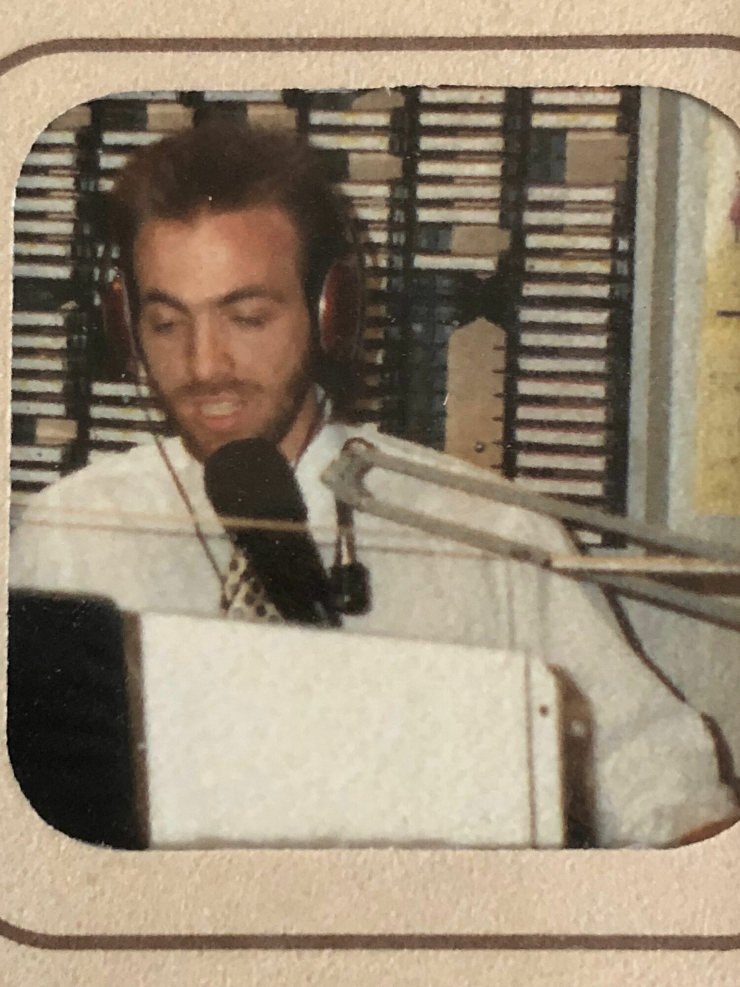 Person speaking into a microphone at a desk with a computer, in a room with shelves and equipment.