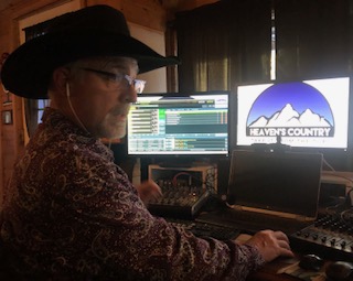 Person wearing a hat and glasses working at a desk with multiple screens, one displaying a mountain logo and text.