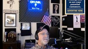 Woman wearing headphones and glasses in a radio studio with a microphone, American flag, and various posters.