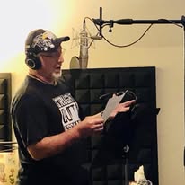 Person wearing headphones and a cap, reading from a script in a recording studio, with a microphone overhead.