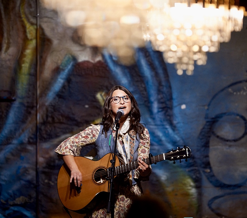 Young woman with glasses singing and playing guitar on stage with a microphone, colorful background, and chandelier overhead.