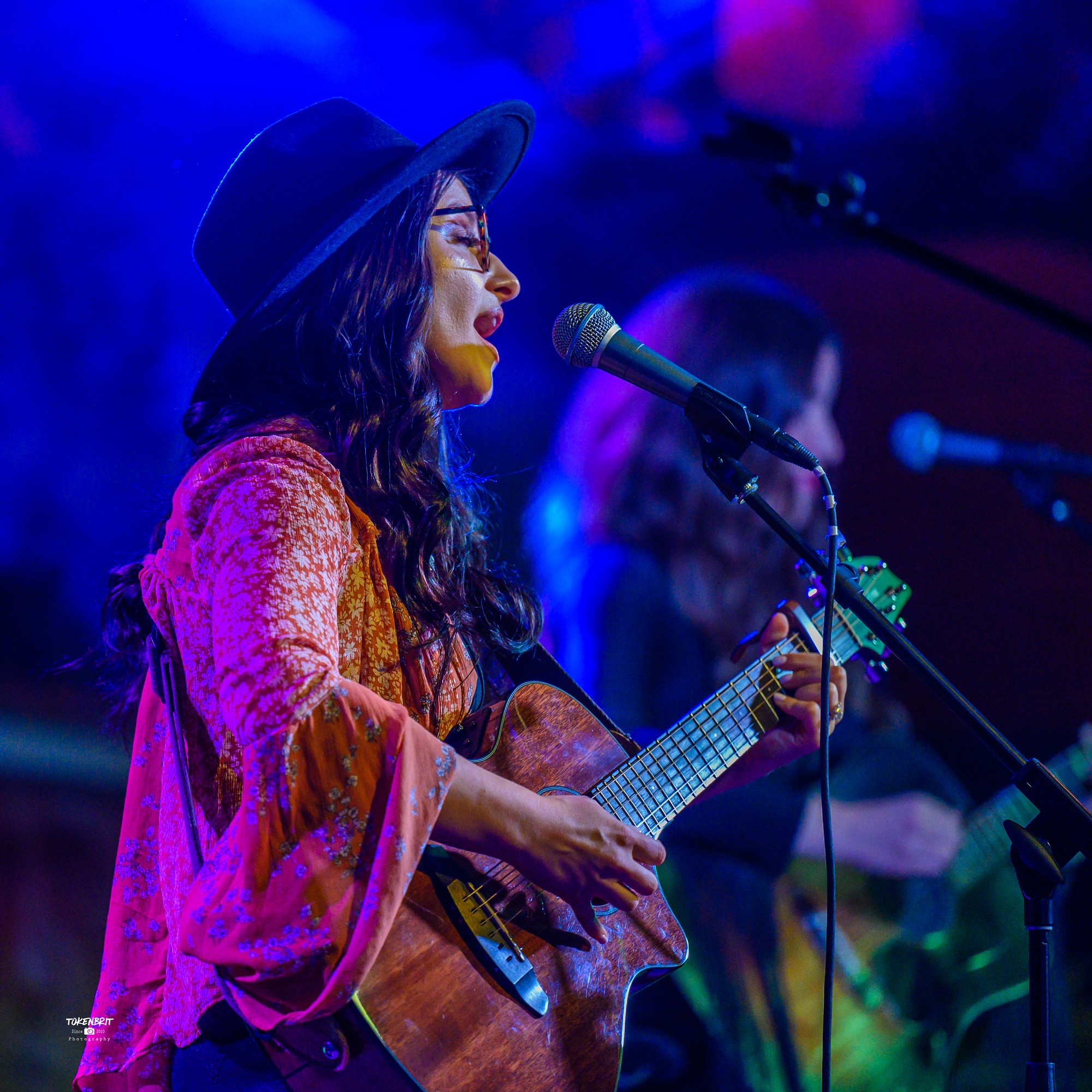 A woman wearing a hat and glasses sings into a microphone while playing an acoustic guitar on stage.