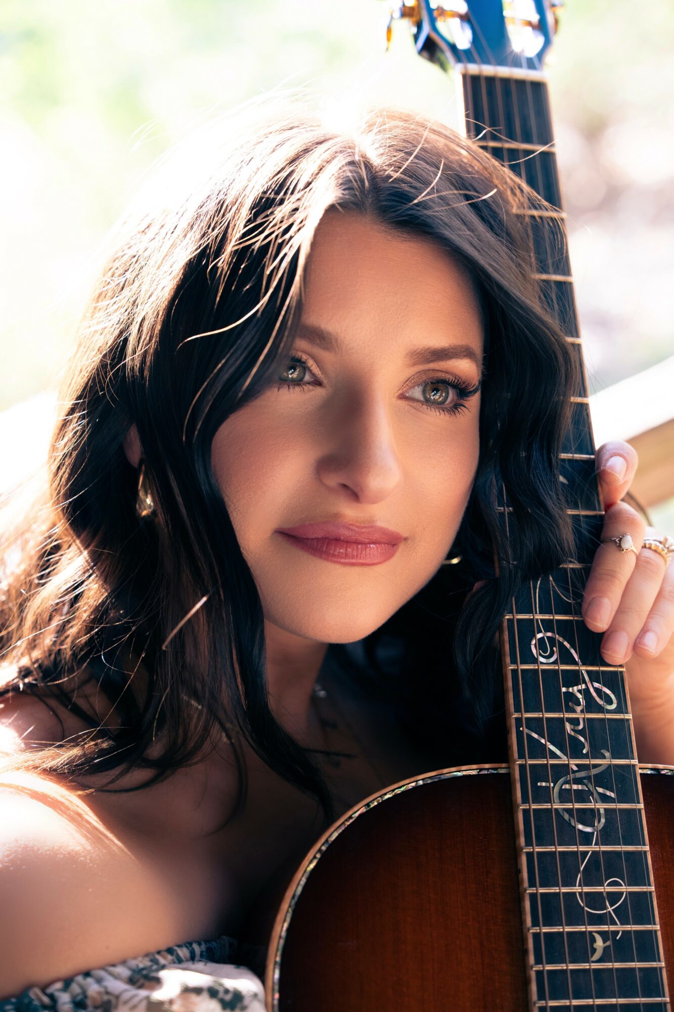 Close-up of a woman with long dark hair holding a guitar, outdoors with blurred background.