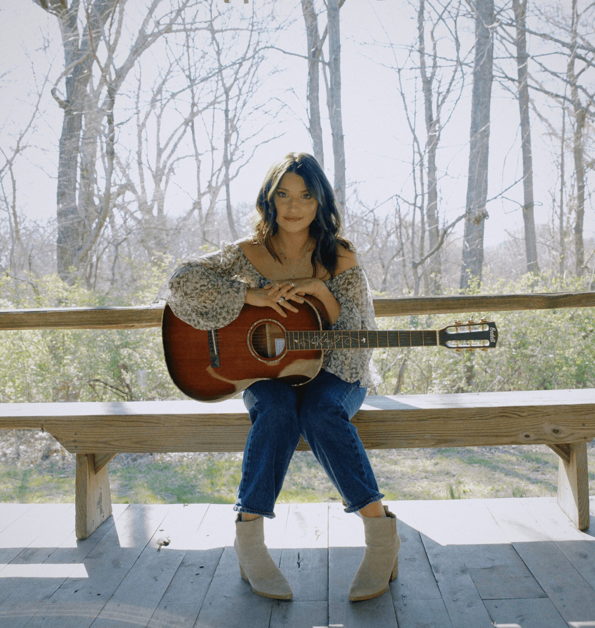 Woman sitting on a wooden bench holding an acoustic guitar outdoors with trees in the background.