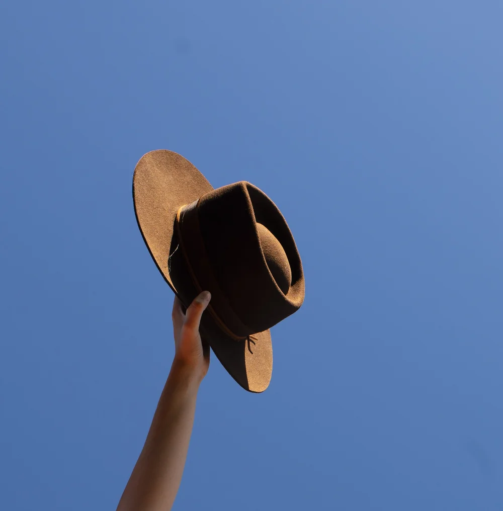 Person holding a brown hat against a clear blue sky, arm extended upward.