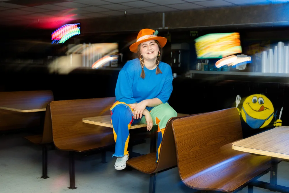 Child wearing a blue outfit and orange hat sitting on a bench in a room with blurred colorful lights, smiling, with a smiley face bag nearby.