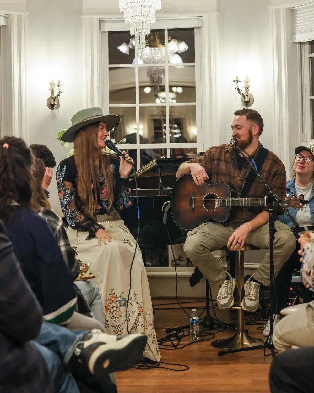 Two musicians perform with a woman singing into a microphone, surrounded by seated audience in a decorated room with chandeliers.