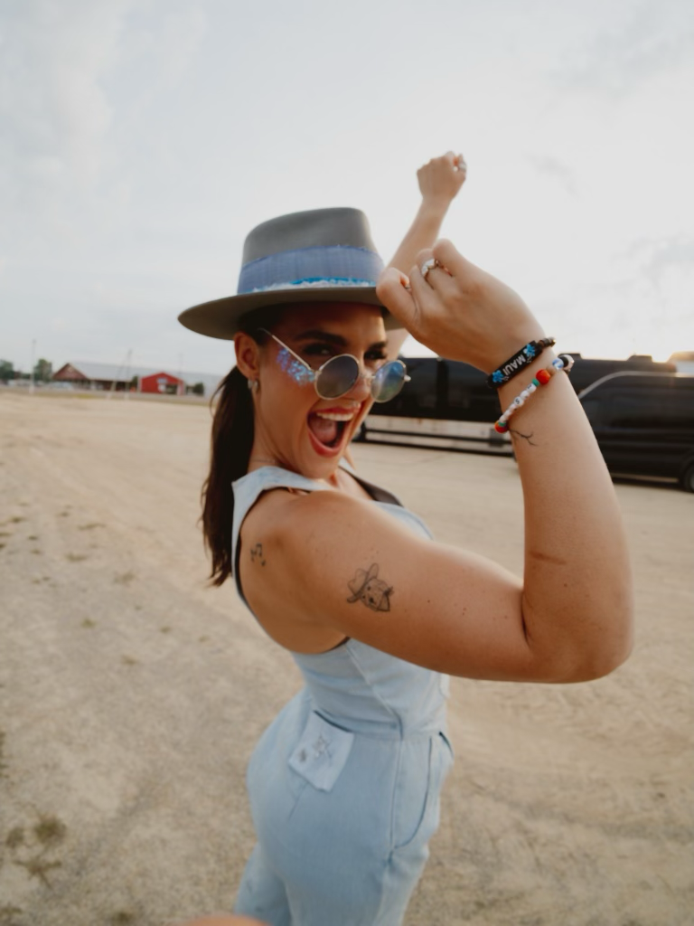 Woman in sunglasses and hat smiling, flexing arm, outdoors on sandy ground, wearing light blue outfit, with a tattoo on her arm.