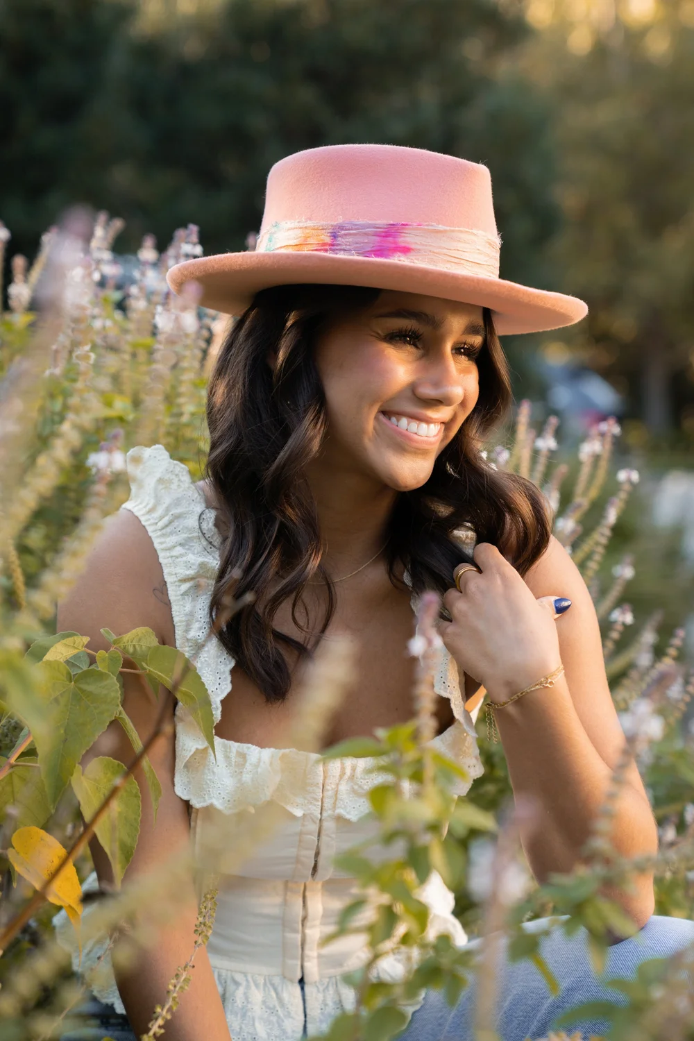 Smiling woman wearing a pink hat outdoors among greenery and flowers, sunlight on her face.