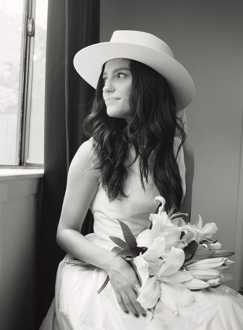 Woman wearing a wide-brimmed hat holding a bouquet of flowers, sitting by a window, looking to the side.