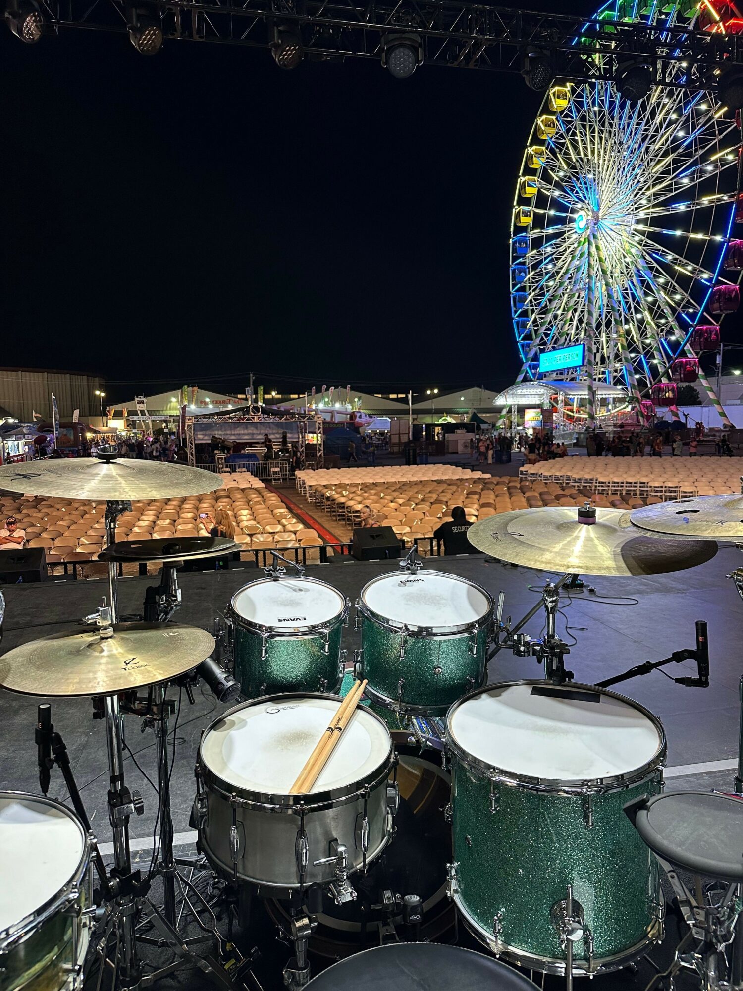 Night scene with a lit Ferris wheel, food stalls, and a drum set on stage at an outdoor event.