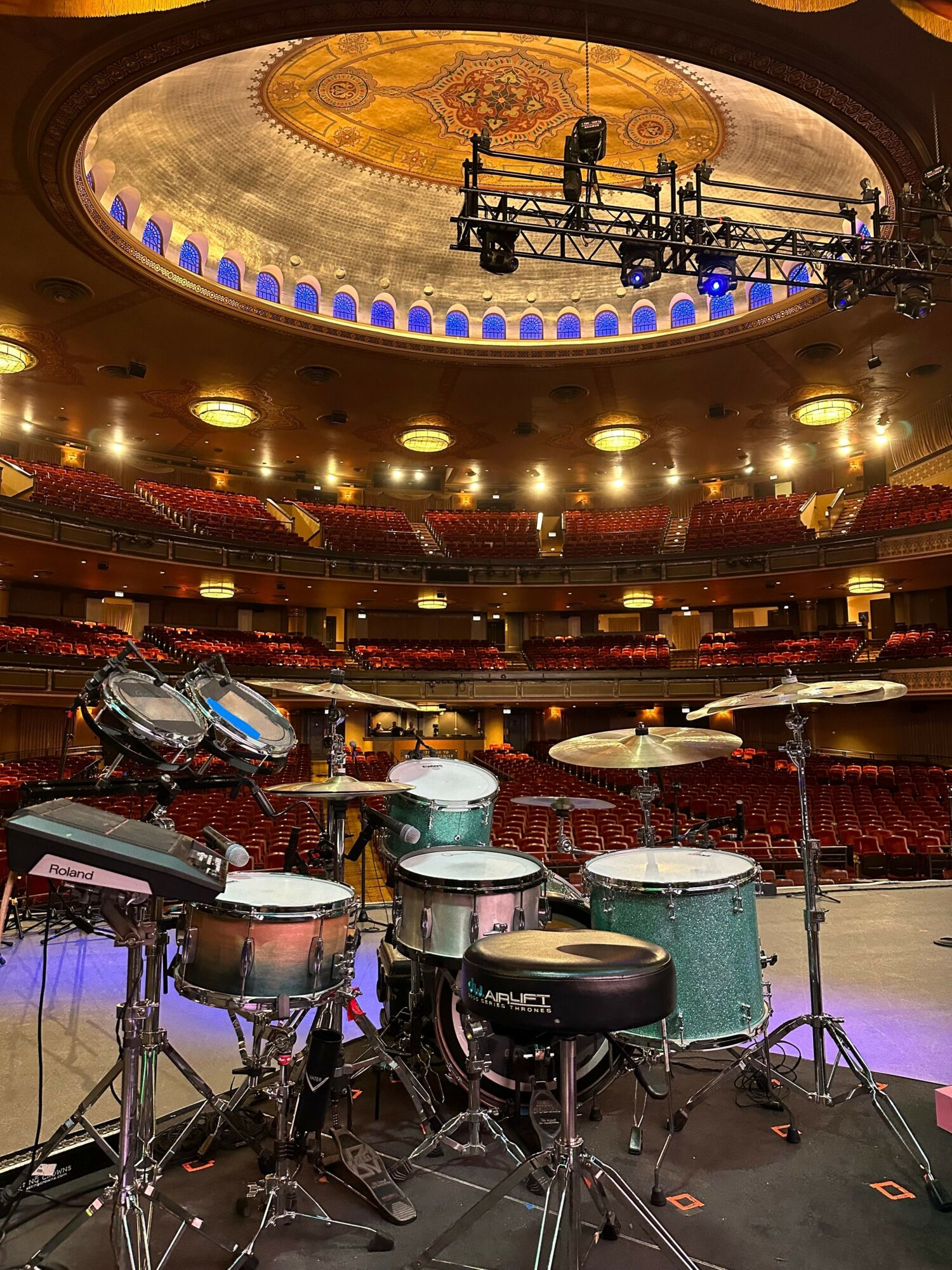 Stage with musical instruments in an empty theater, audience seats visible, stage lights overhead, and a drum set on the right.
