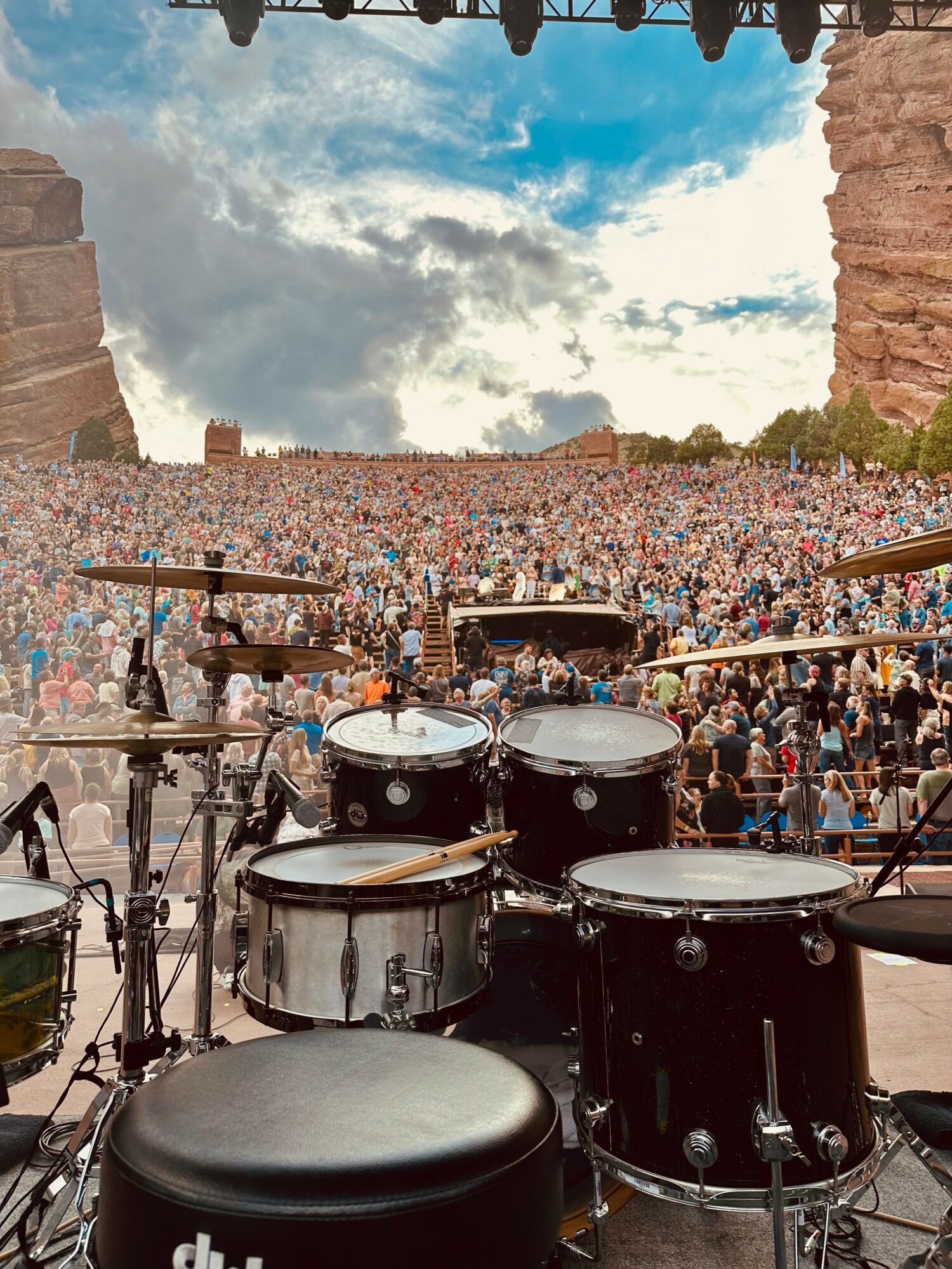 Stage with drums facing a large crowd outdoors, sky with clouds overhead, stone structures on sides, audience in distance.