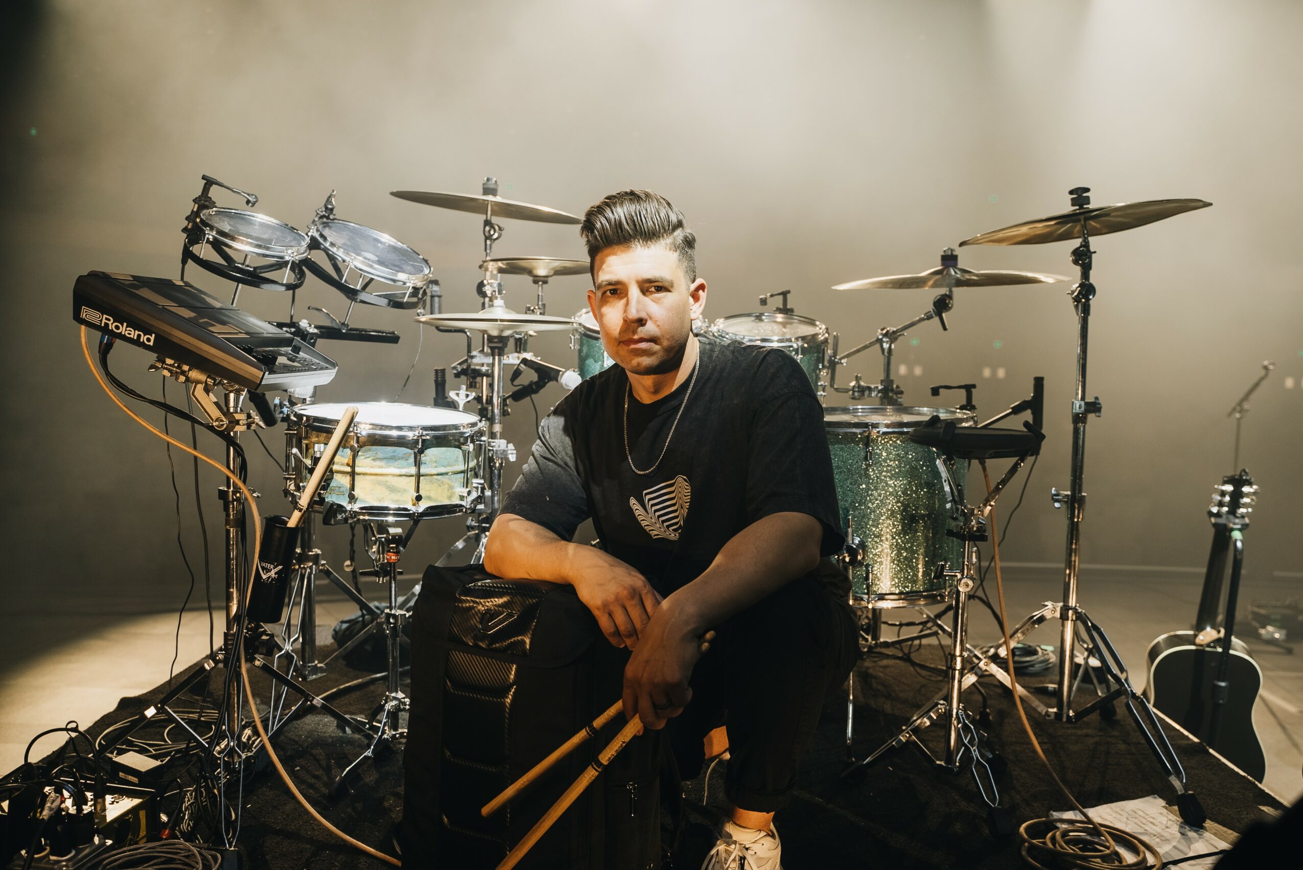 Man sitting in front of a drum set on stage, holding drumsticks, with musical equipment around him.