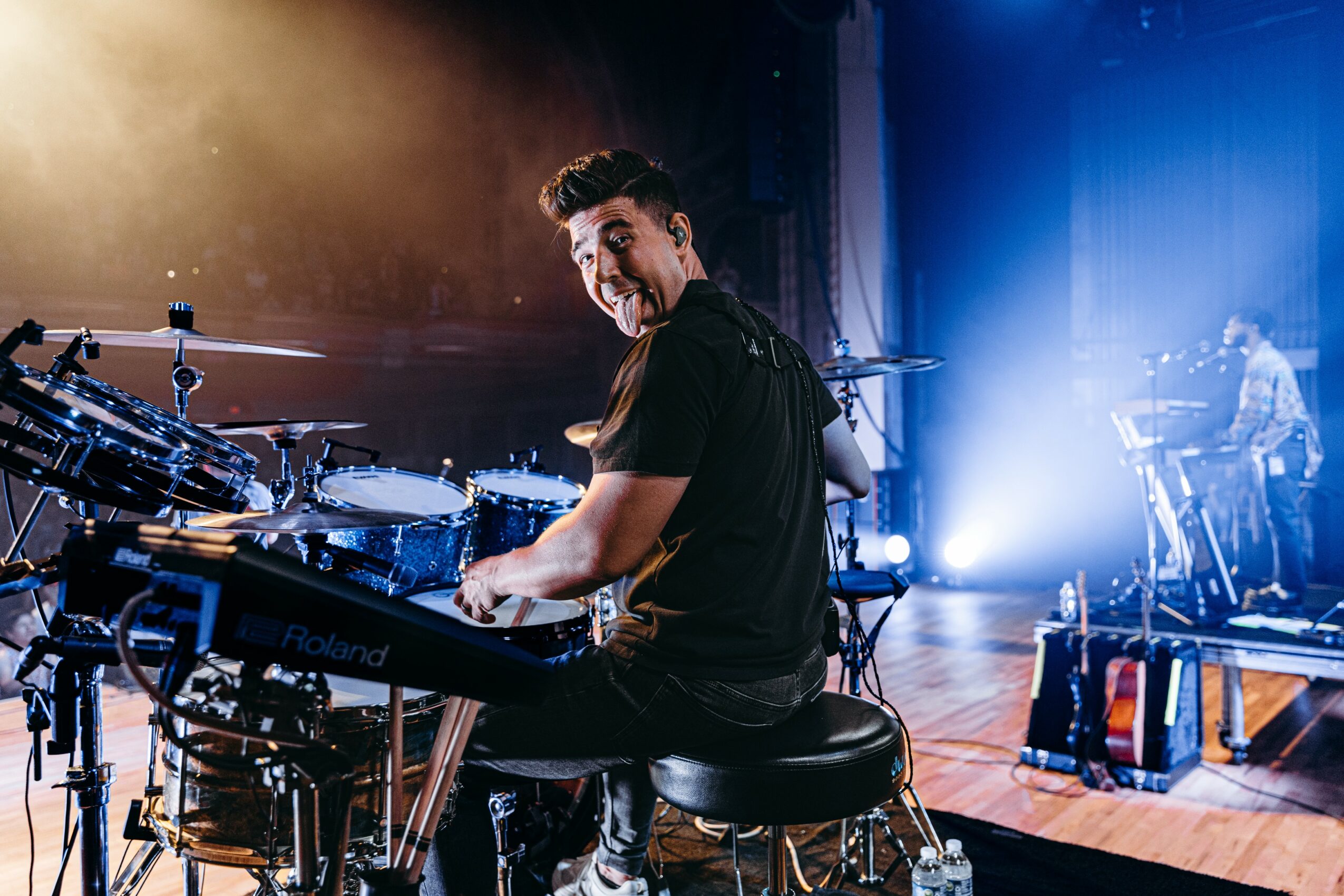 Musician playing keyboard on stage with drums and electronic equipment, smiling, stage lighting in background