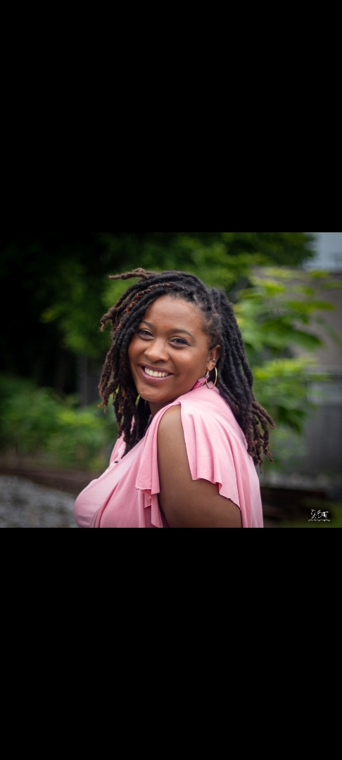 Smiling woman with curly hair wearing a pink top outdoors with greenery in background.