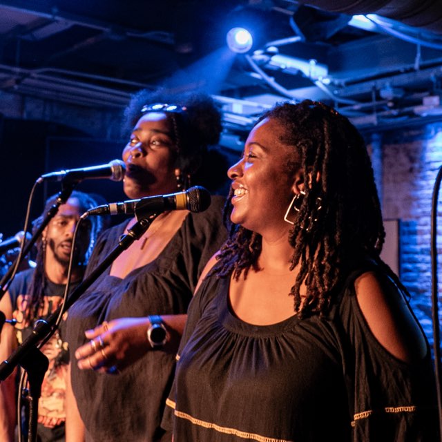Two women singing into microphones on stage, smiling, with a man in the background, in a dimly lit venue.