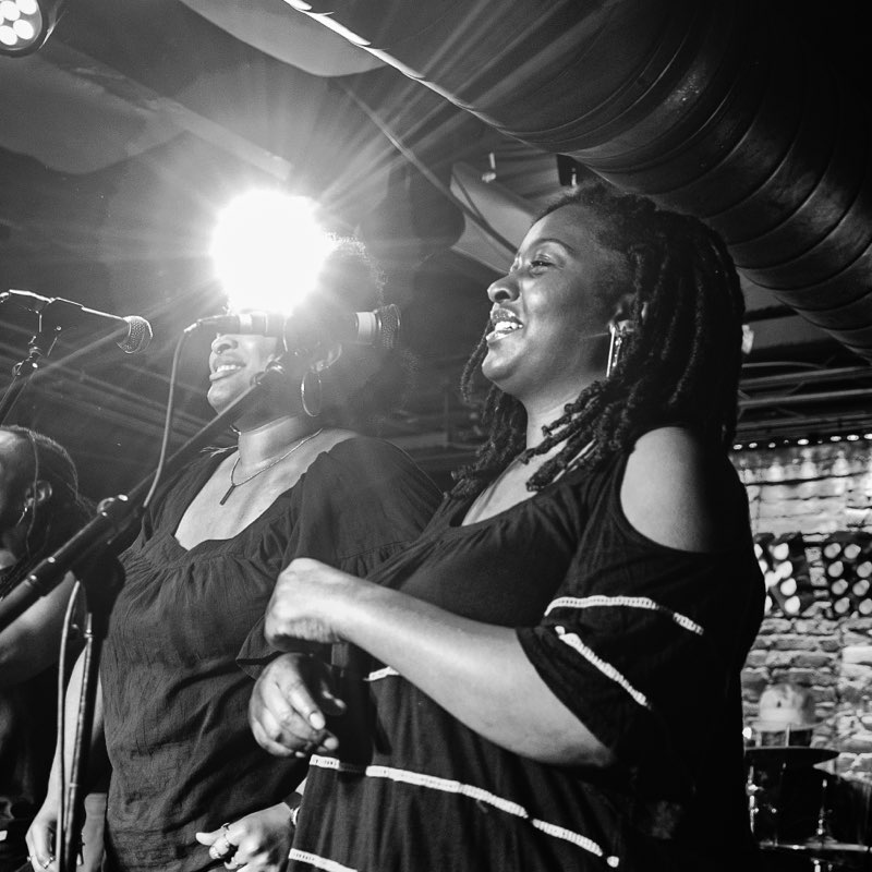Two women singing on stage, one smiling with dreadlocks, bright light behind them, in a lively performance setting.