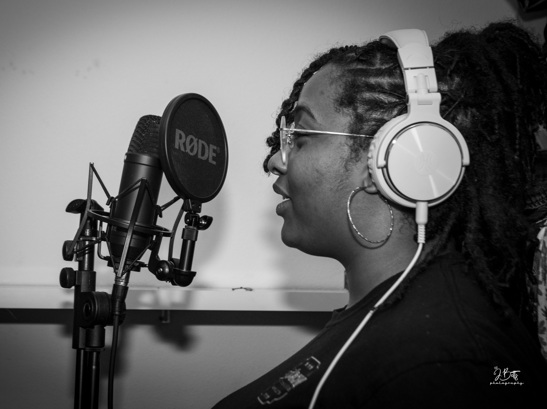 Woman with dreadlocks and glasses wearing headphones, speaking into a microphone in a recording studio.
