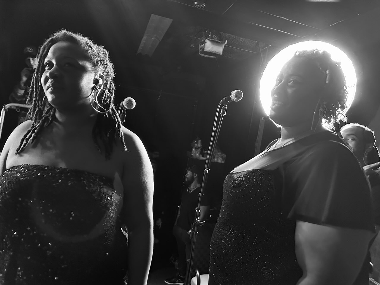 Two women stand on stage with microphones, one with dreadlocks and the other with curly hair, illuminated by stage lights.