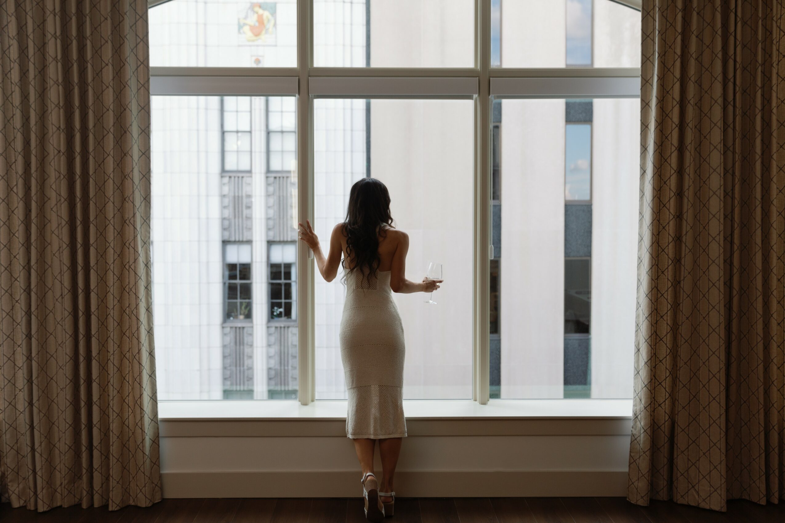 Woman in a white dress looking out a large window with city buildings outside, curtains on sides, natural light.