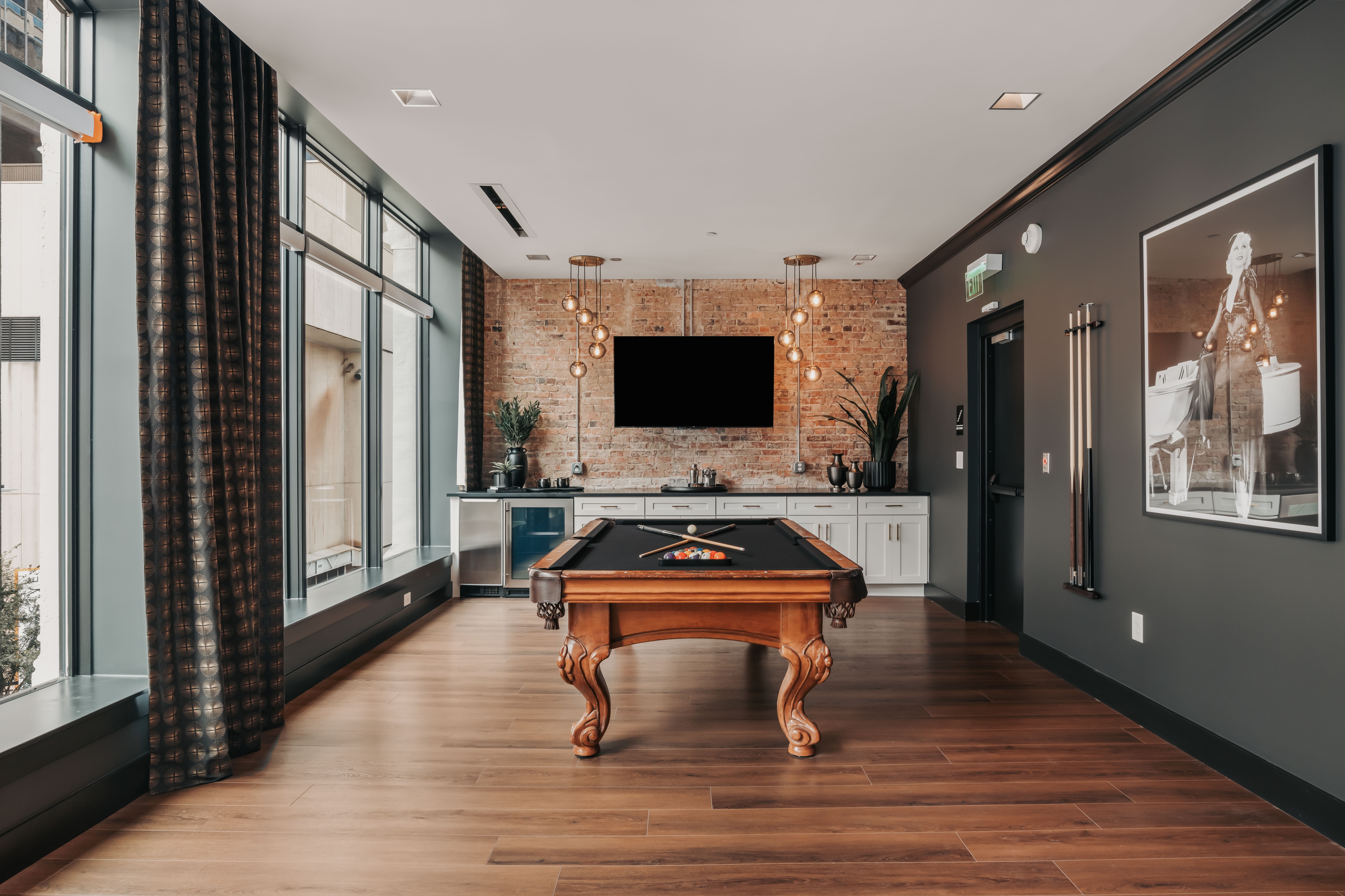 Room with a pool table, large windows, a TV on brick wall, and modern decor, including plants and artwork.