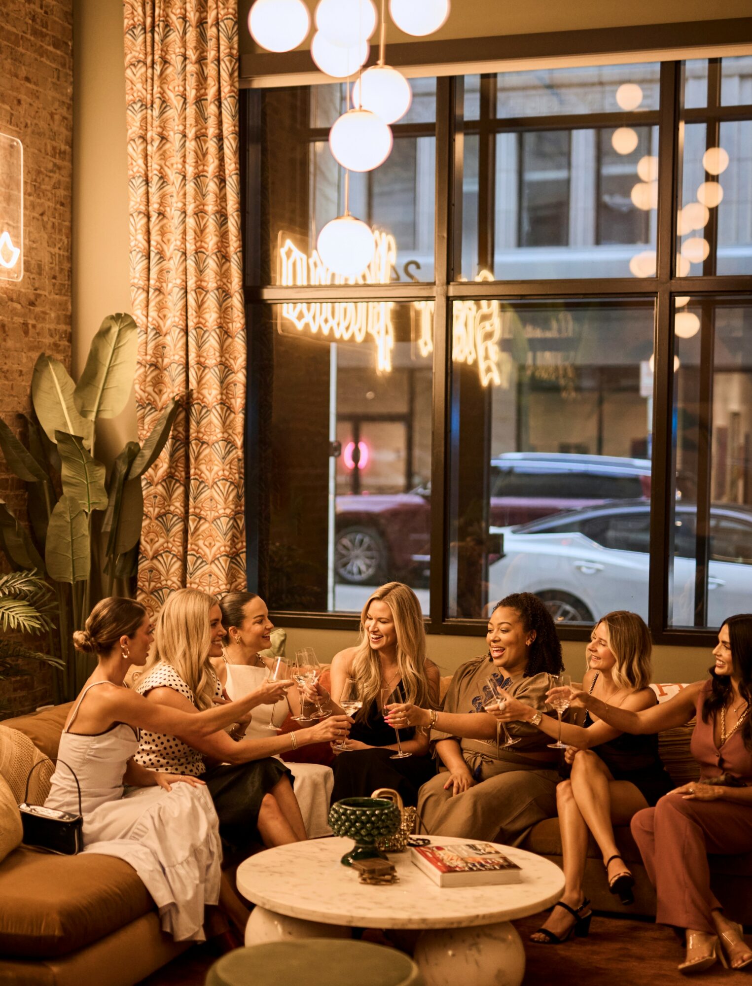 Group of women sitting on a couch, reaching out to each other, in a warmly lit room with large window and plants.