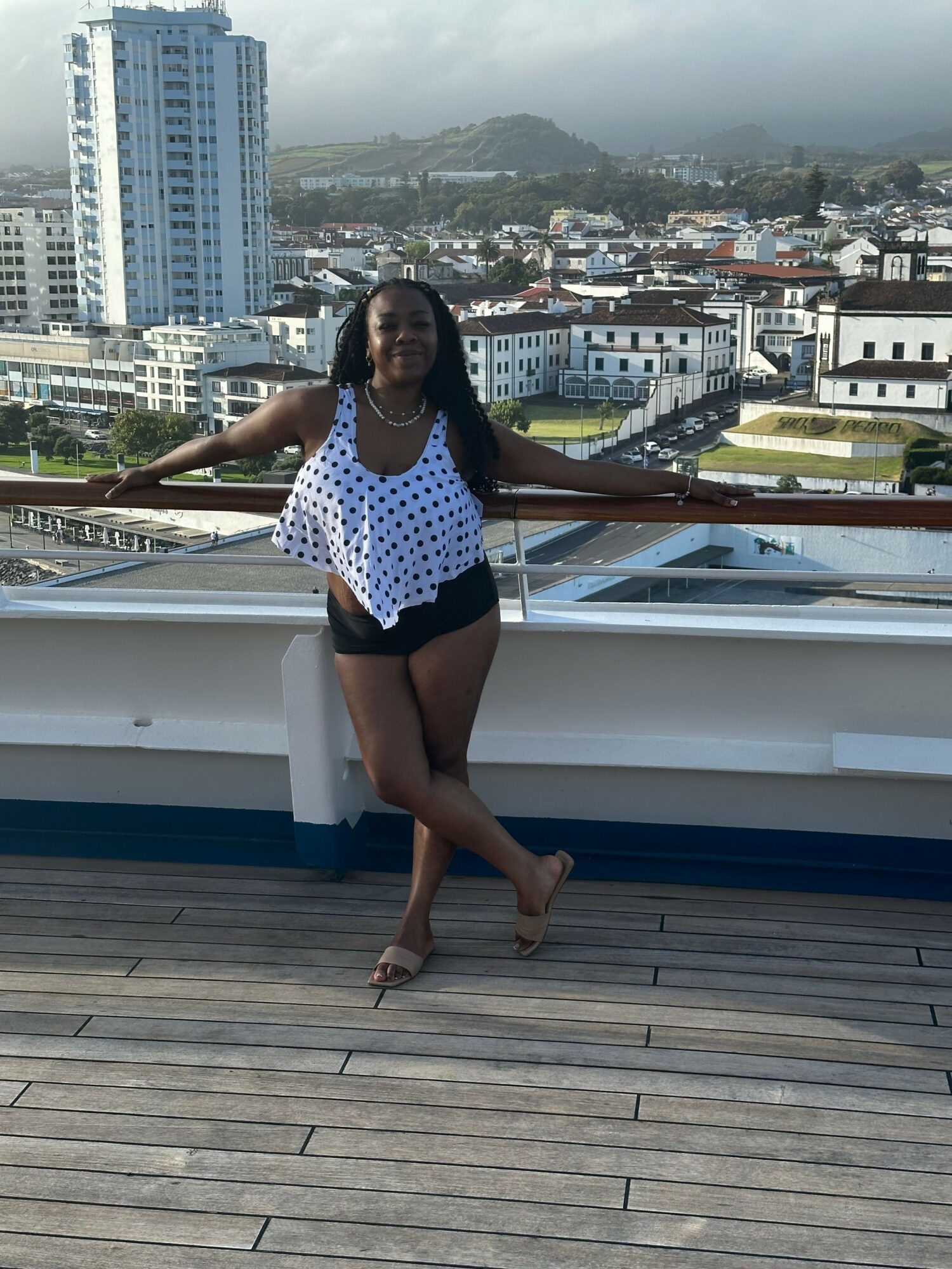 Woman in polka dot top and shorts standing on a boat deck with cityscape background.