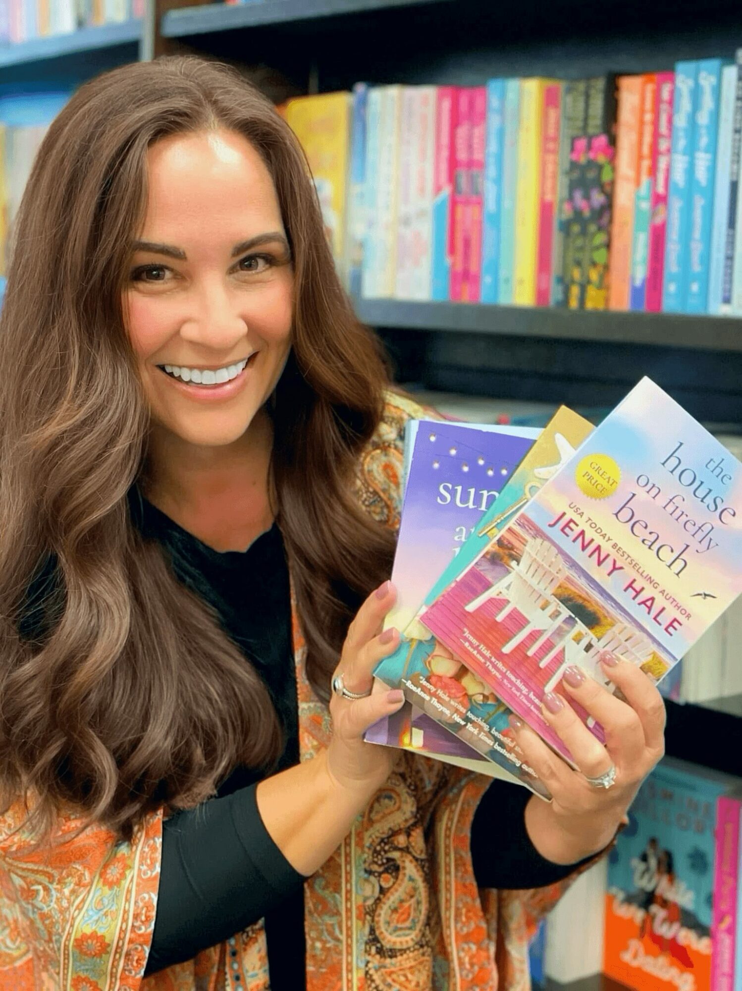 Woman with long brown hair smiling, holding a stack of colorful books in a bookstore or library.