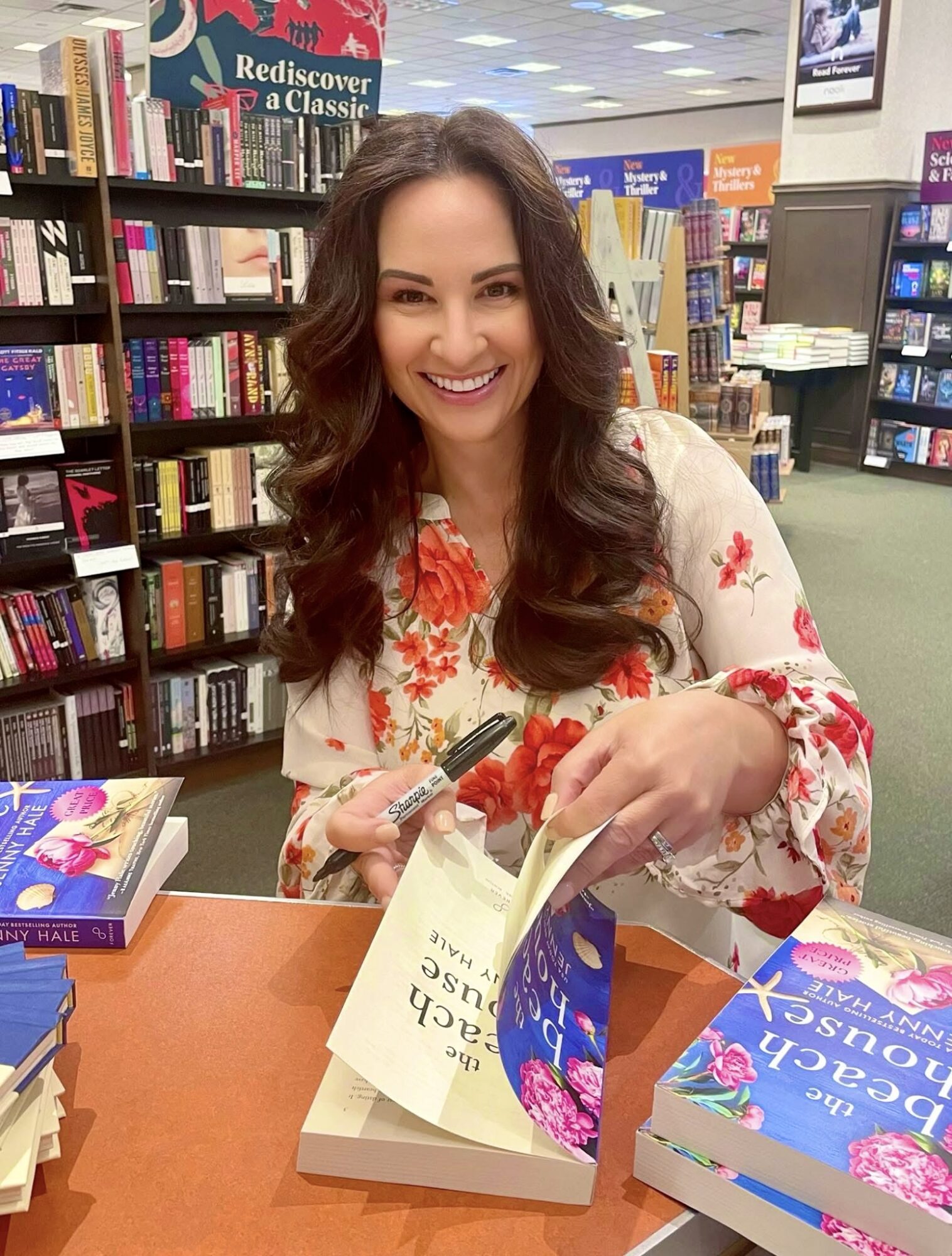 Woman with long wavy hair smiling at a book signing event, surrounded by books in a bookstore.