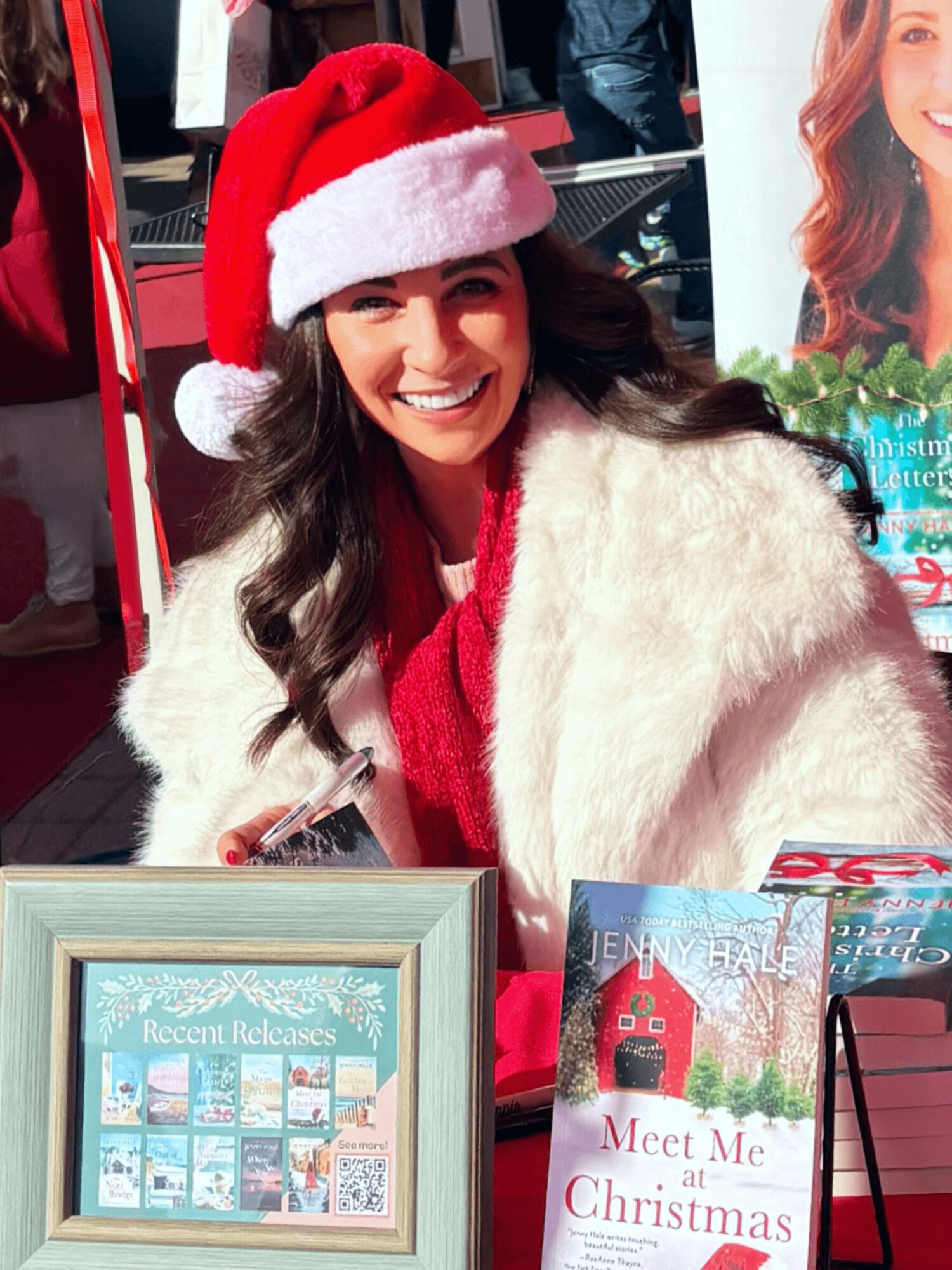 Woman wearing a Santa hat and white fur coat smiling at a Christmas event, with books and a framed display in front.