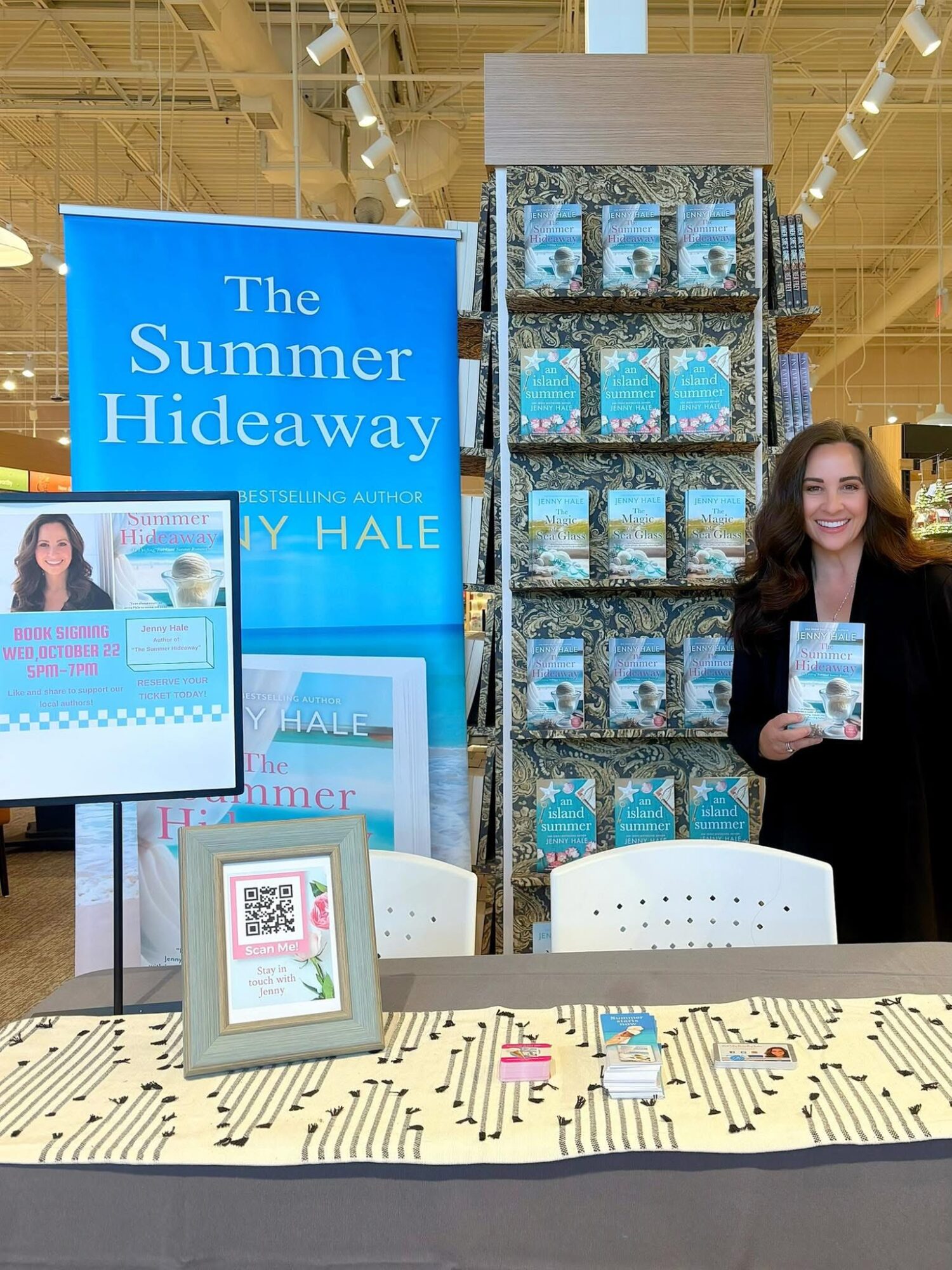 A woman standing next to a display of books and a large blue sign for 'The Summer Hideaway' at a book event.