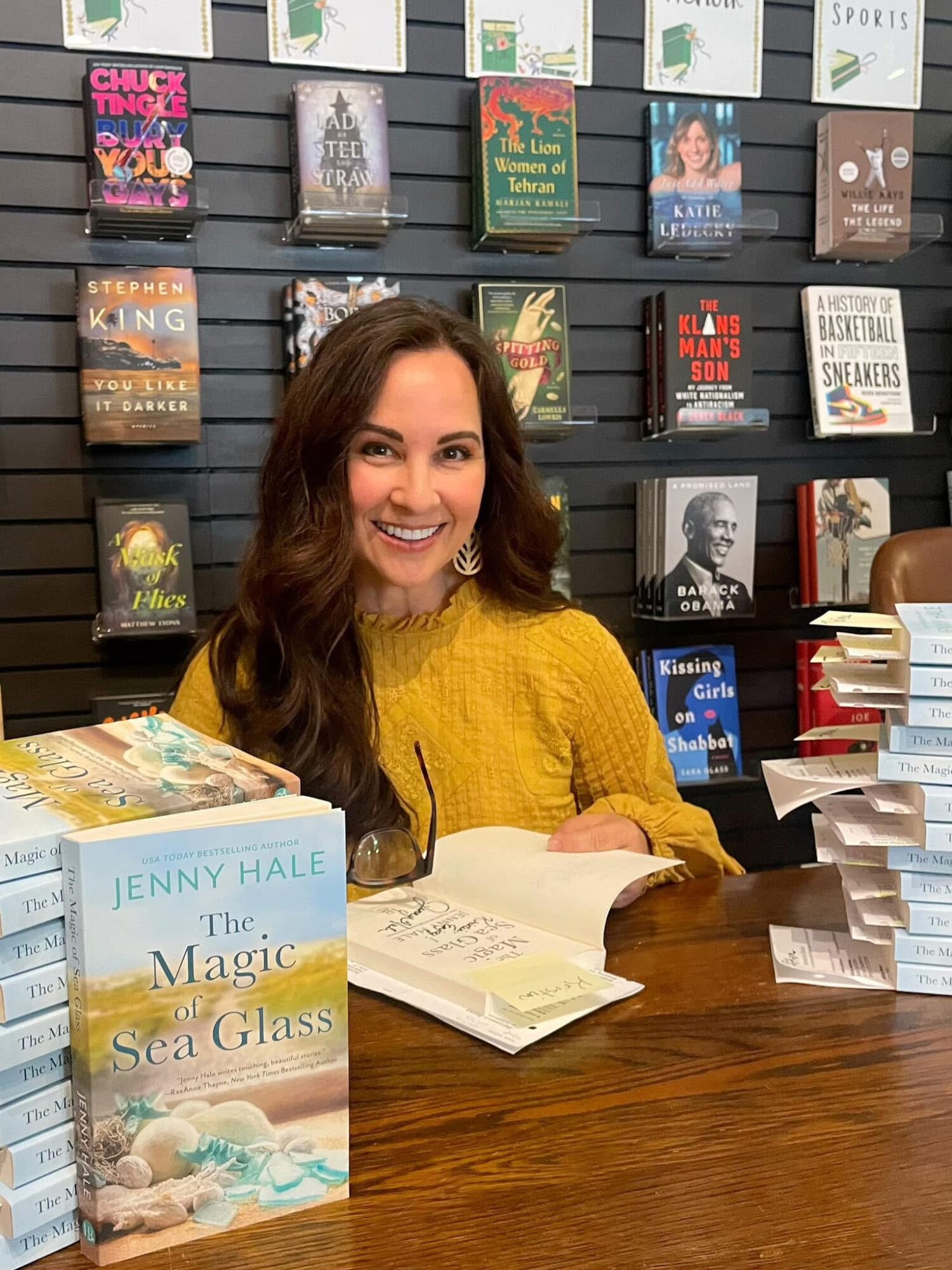 Woman with long brown hair in a yellow sweater sitting at a table with books, smiling, bookshelves in background.
