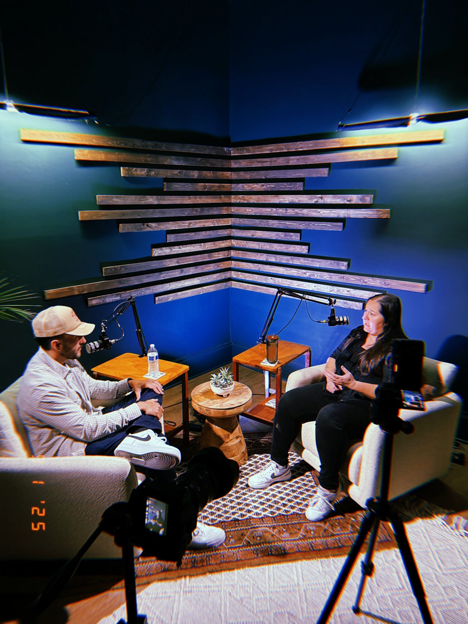 Two people sit in a recording studio with blue walls and wooden acoustic panels, microphones, and recording equipment, engaged in conversation.