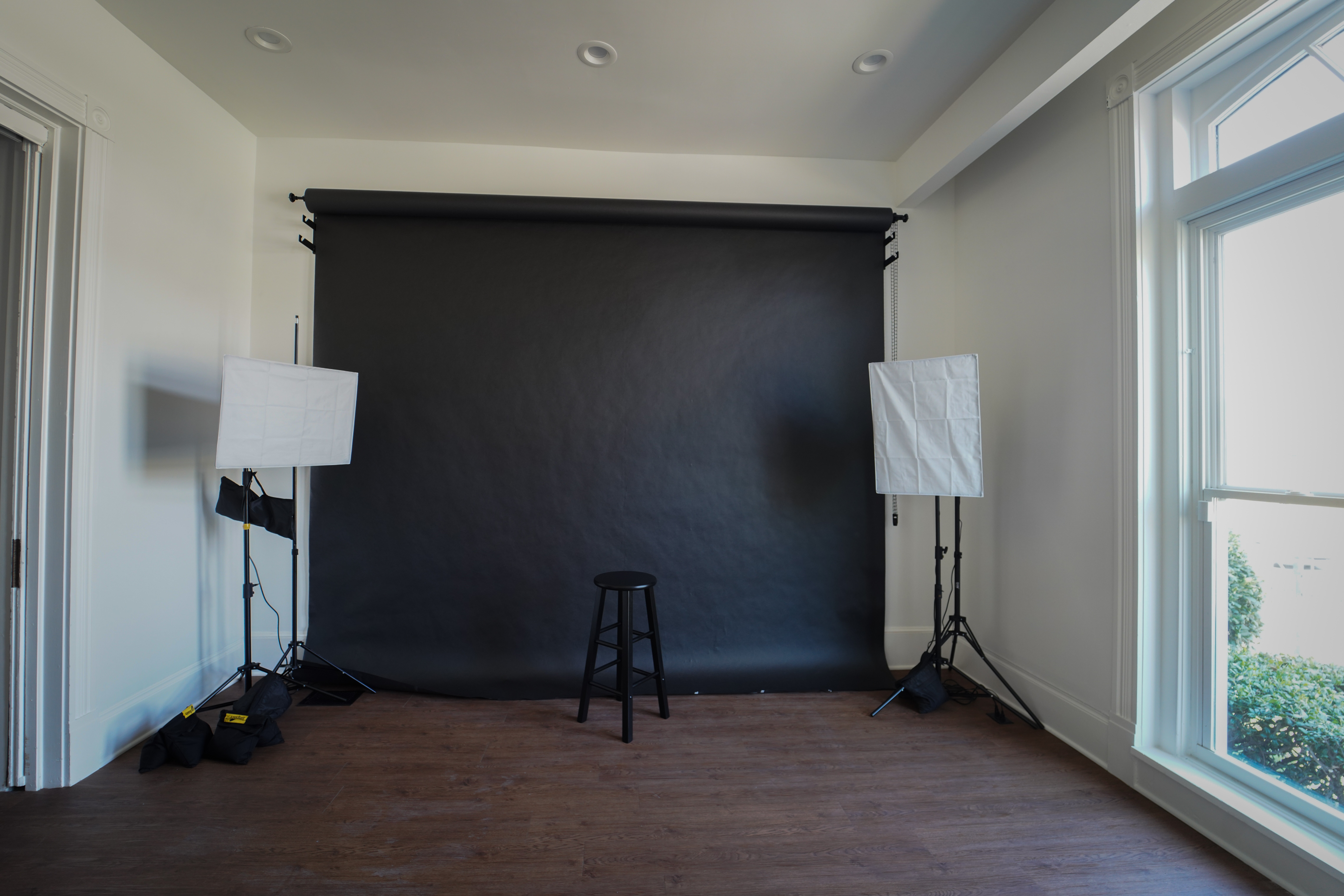 Room with black backdrop, two standing lights, a stool, and large window, hardwood floor, and white walls.