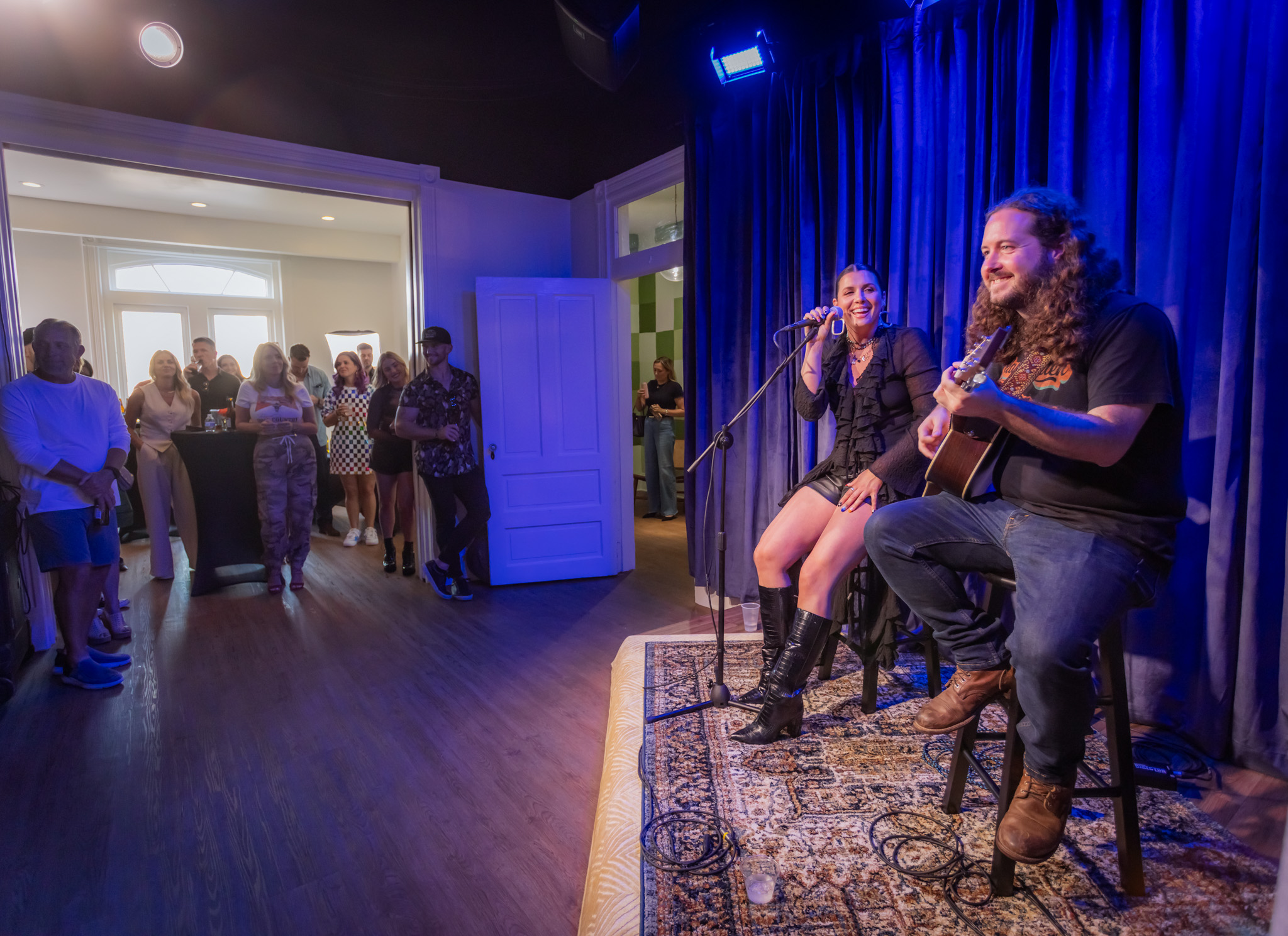 Two performers, a woman and a man, sing and play guitar on a stage with blue curtains, audience in background.