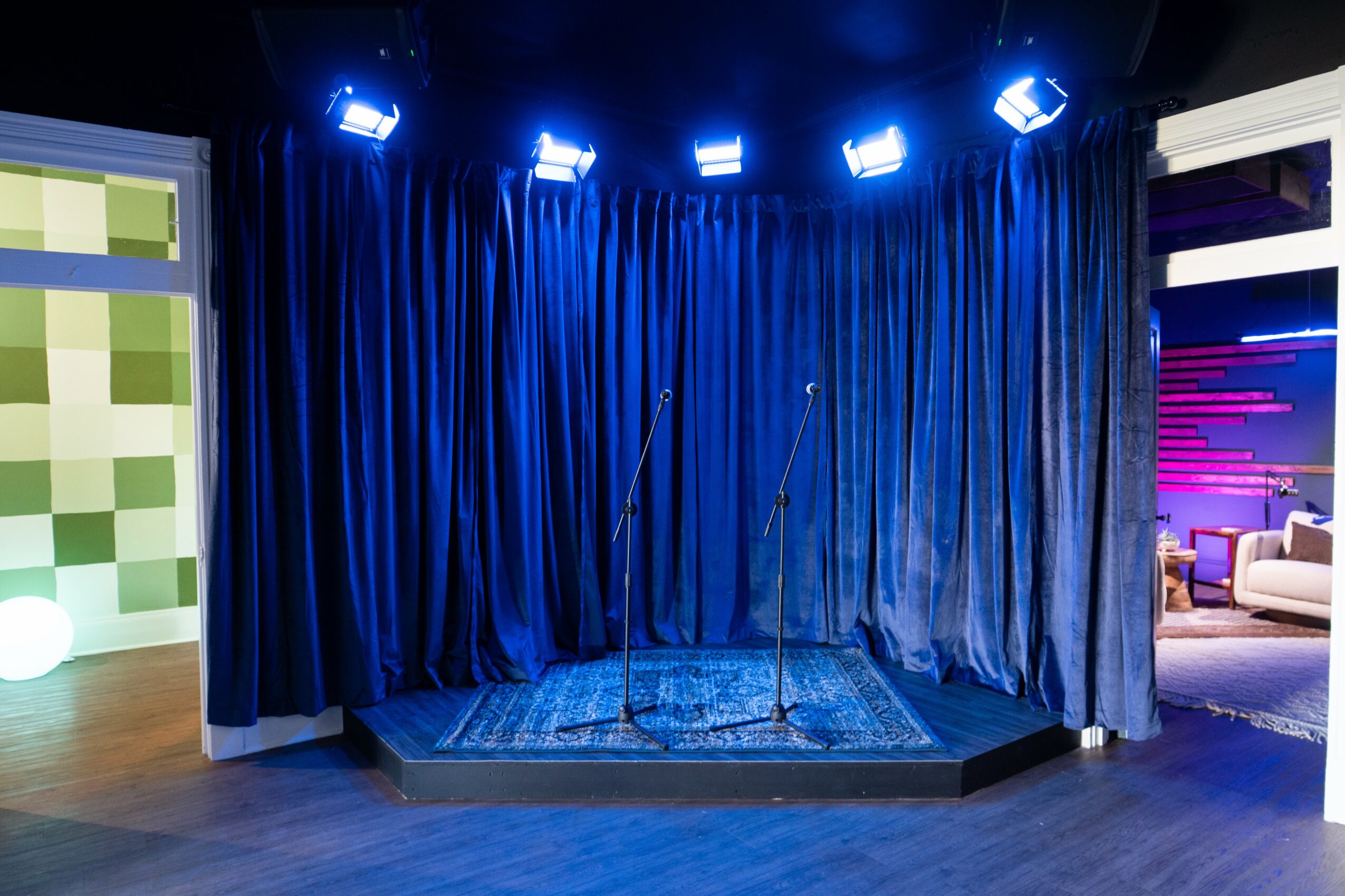 Empty stage with blue curtains and microphone stands, illuminated by stage lights, in a theater or performance space.