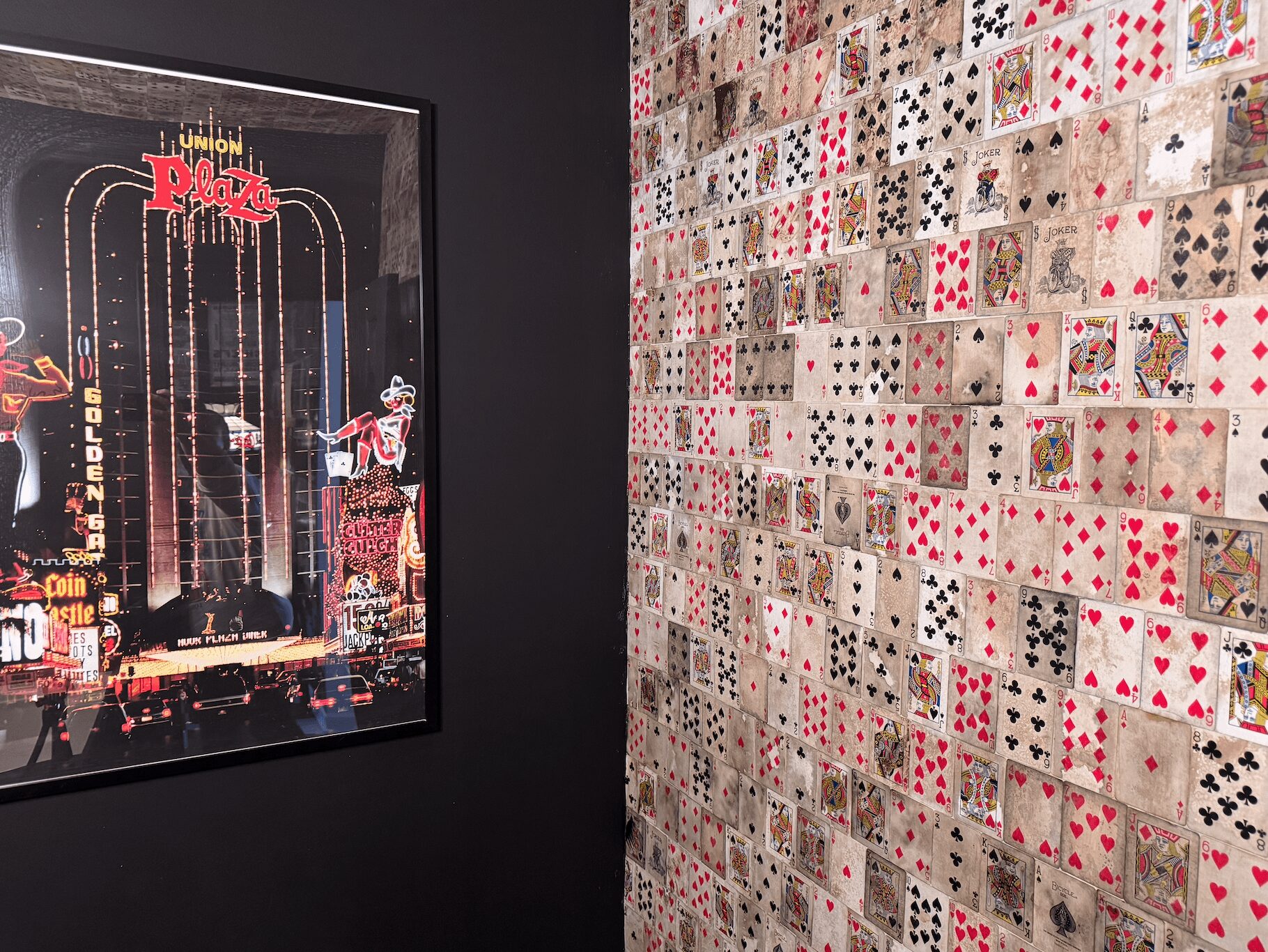 A wall covered with playing cards arranged in a grid pattern, next to a framed poster of a city scene at night.