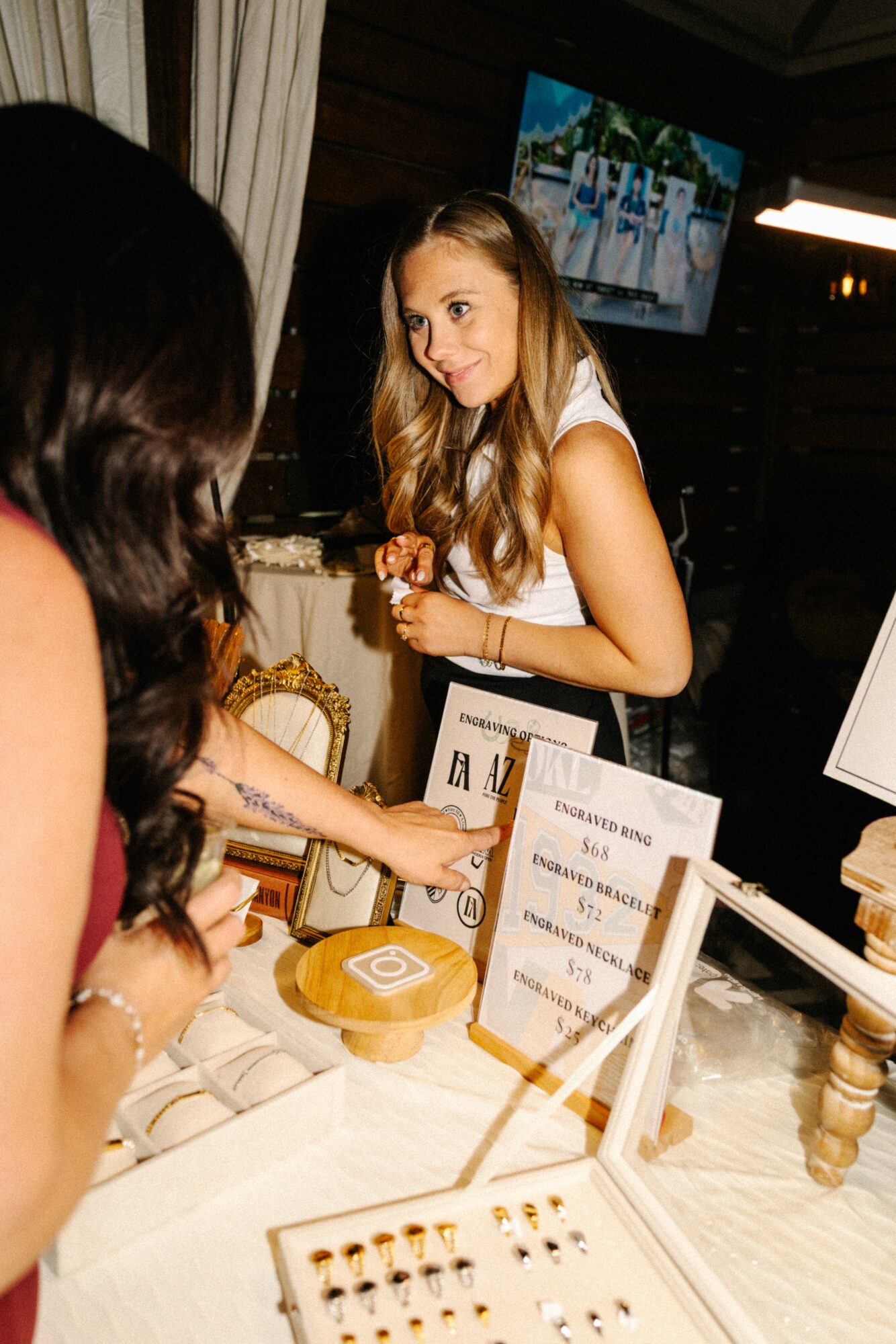 Young woman smiling and talking at a jewelry display table with a person across from her, in a dimly lit indoor setting.